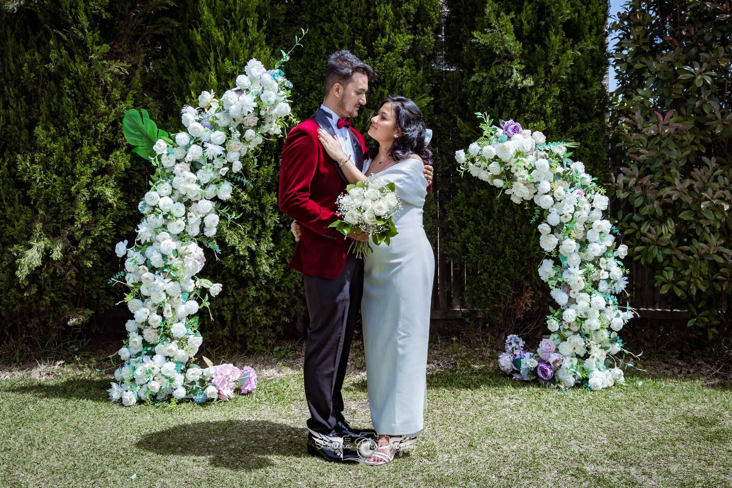 Newlyweds share intimate moment under decorative white floral arch in garden setting.