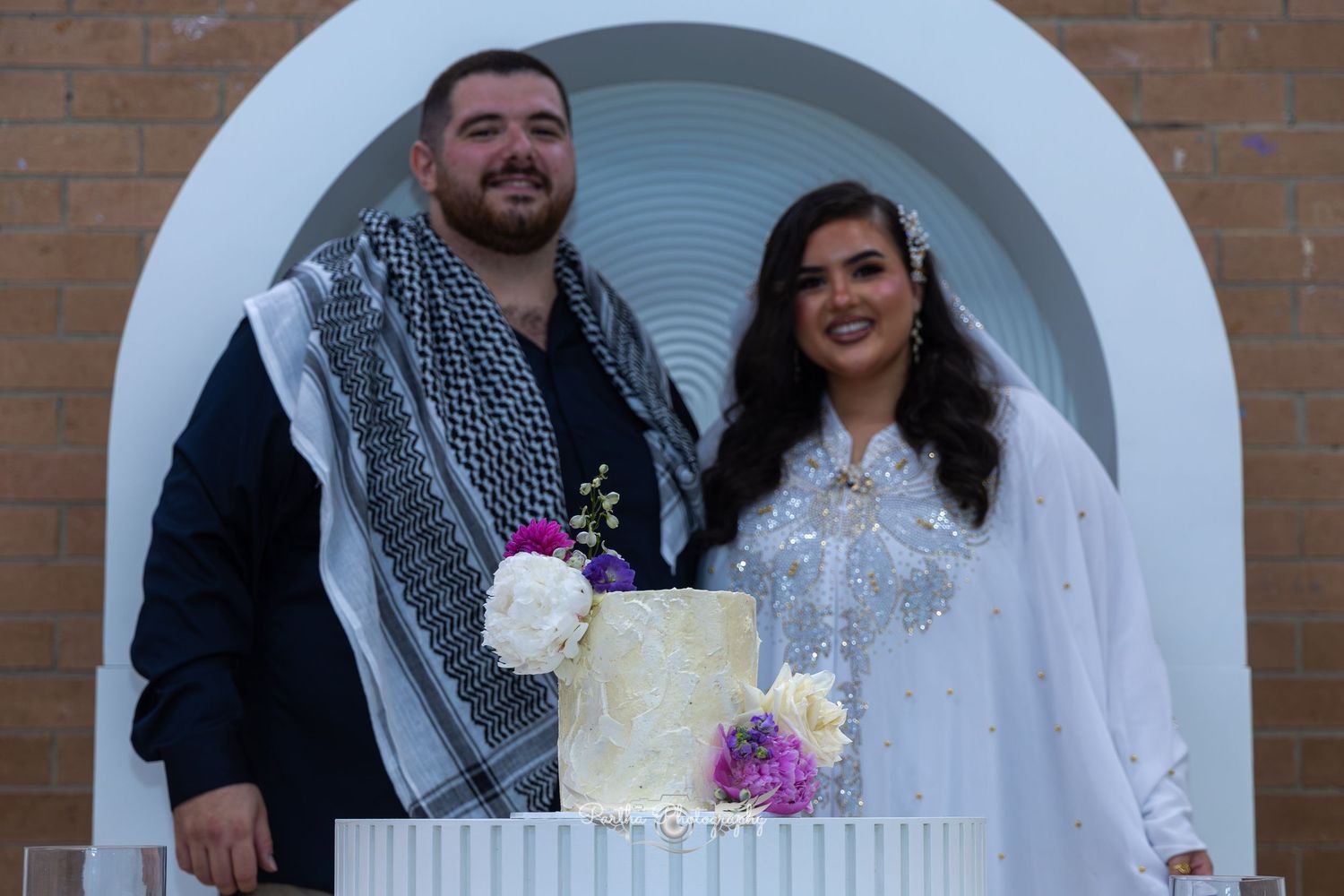 Newlyweds posing with their wedding cake in front of an arched doorway.