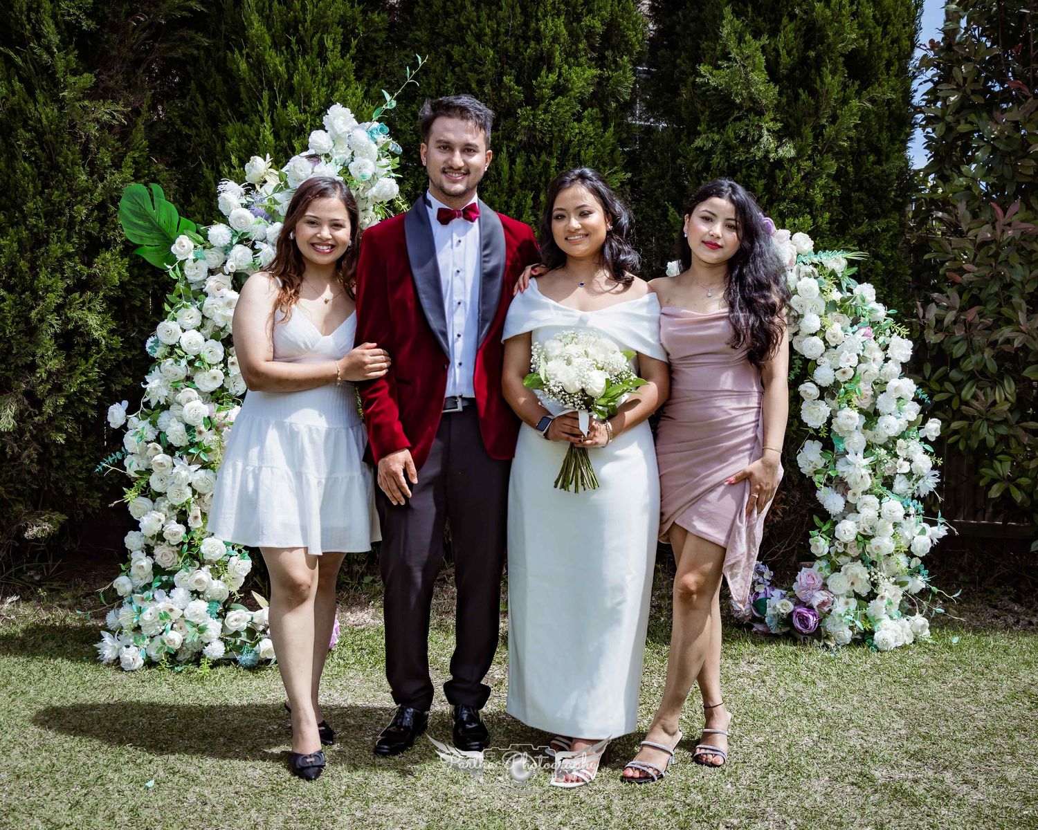 Wedding party of four poses in front of white floral arch during outdoor celebration.