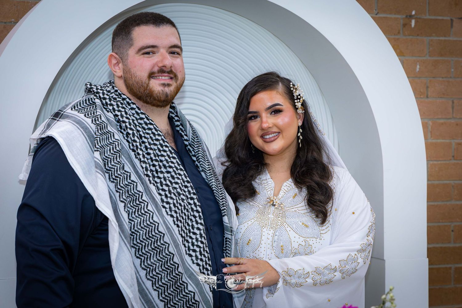 Couple in fusion wedding attire posing formally in front of an arched backdrop.
