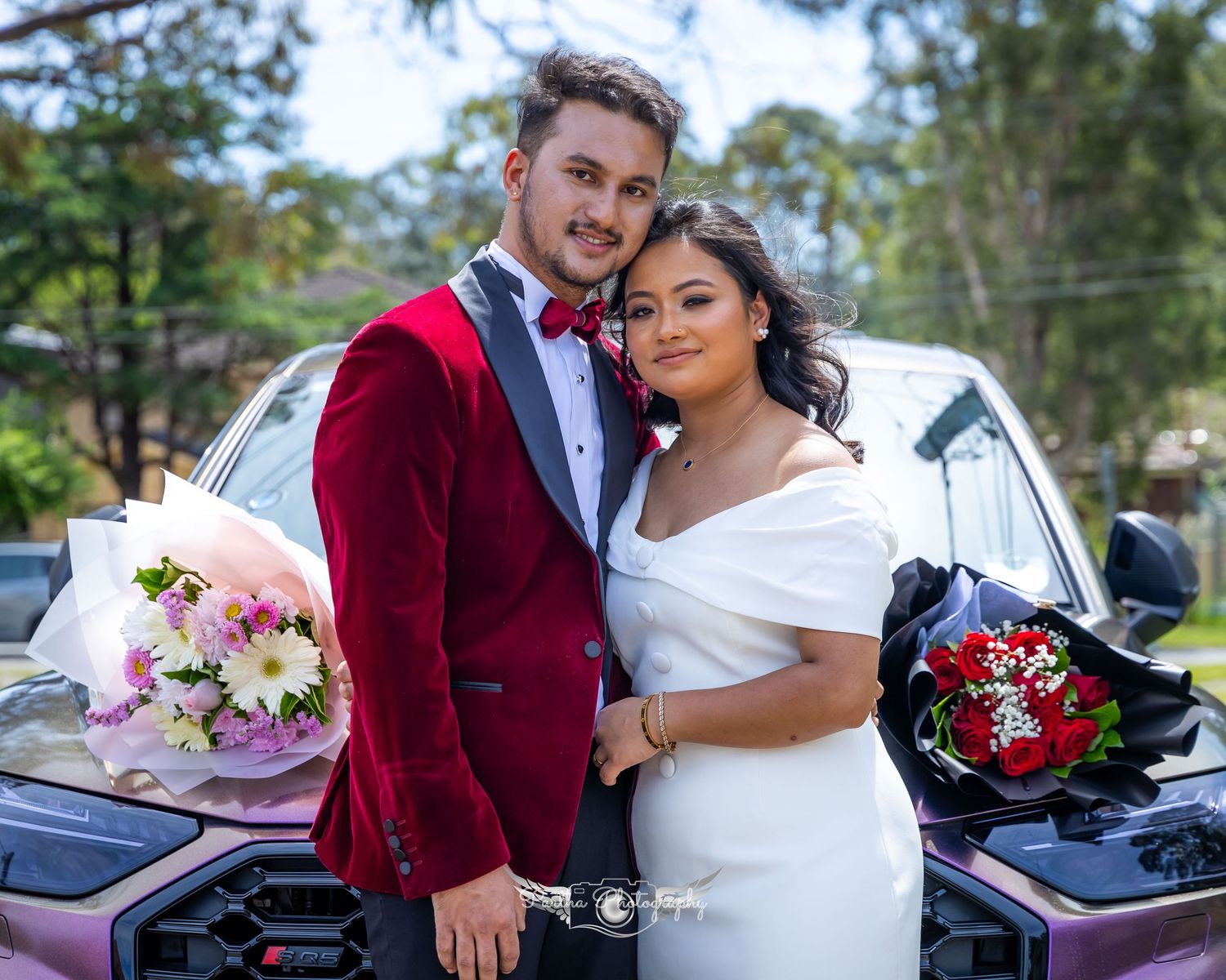 Happy couple poses by car with bouquets, bride in white dress and groom in red velvet jacket.