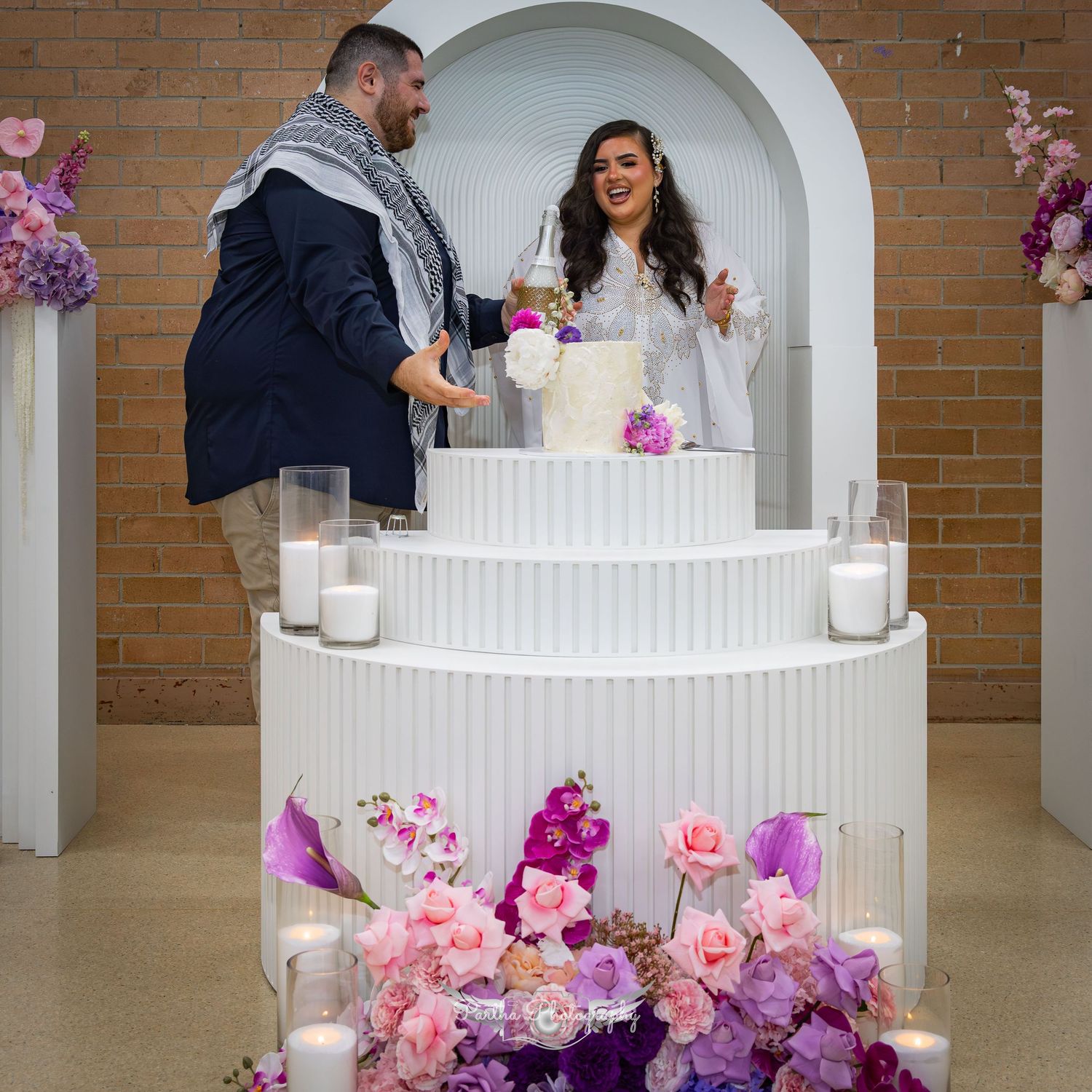 Couple celebrates at white tiered display decorated with pink and purple flowers and candles.