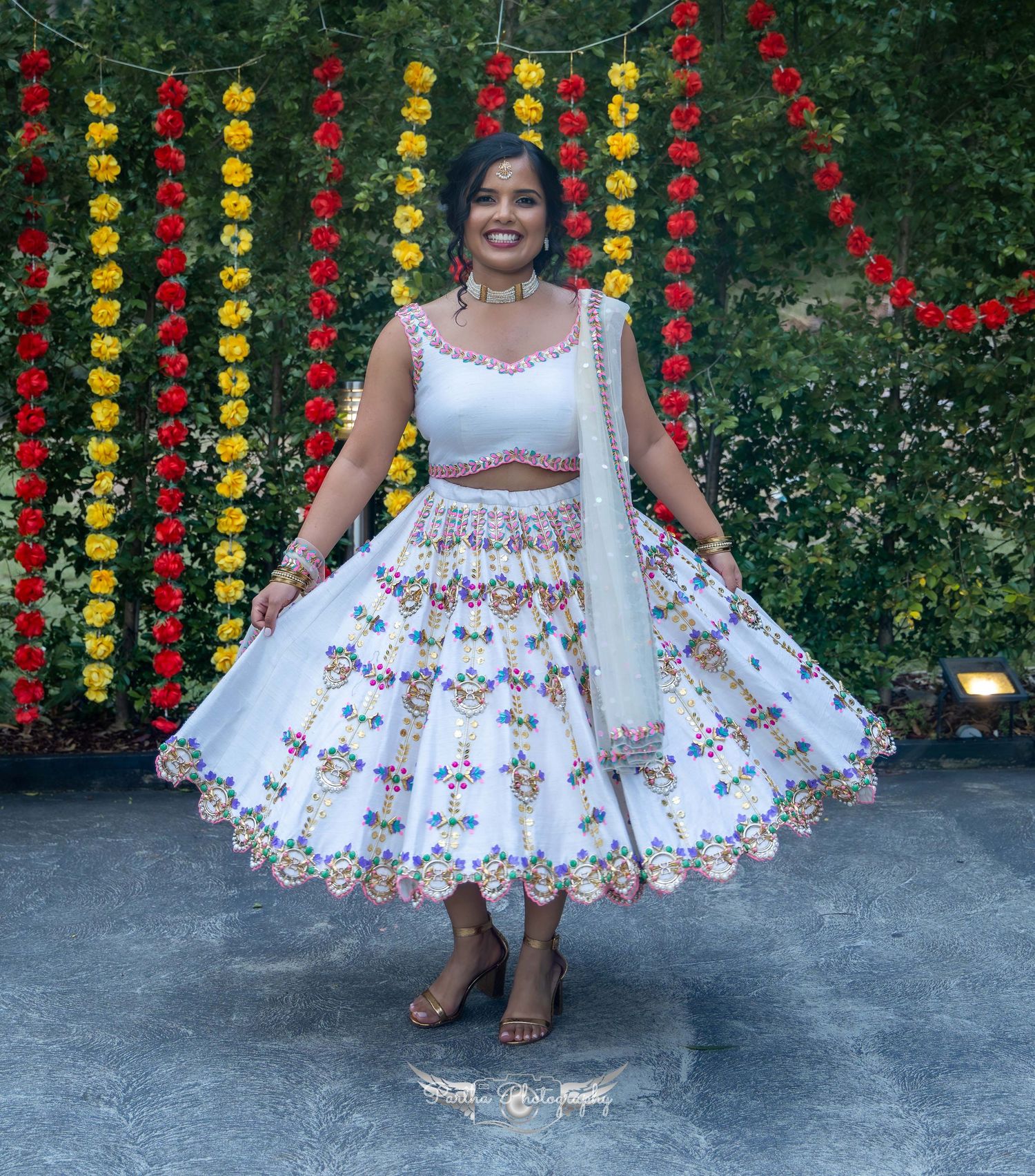 A woman in a white floral lehenga poses in front of colorful paper lantern decorations.
