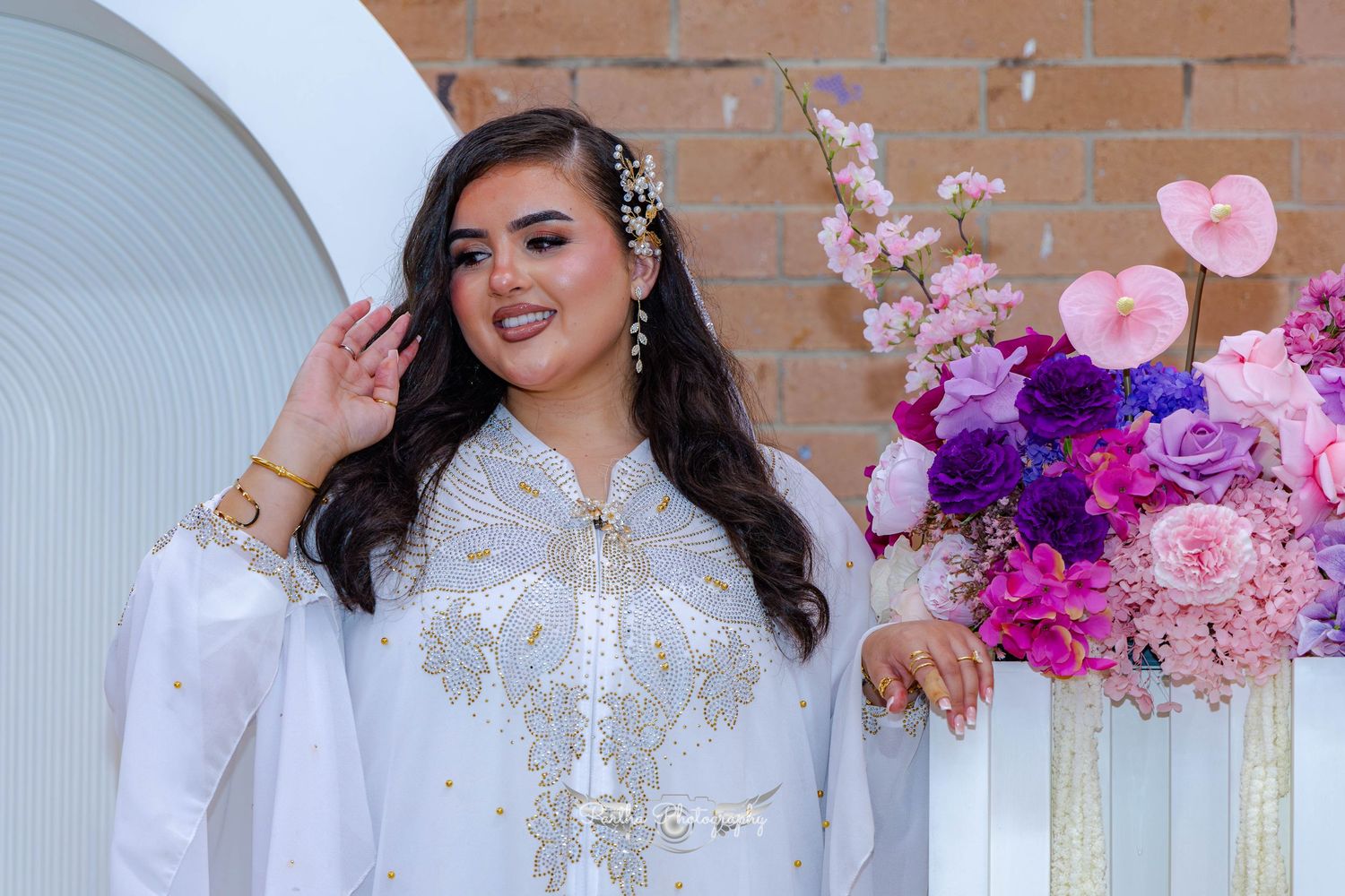 A bride in an ornate white dress poses under a decorated arch with pink and purple flowers and brick backdrop.