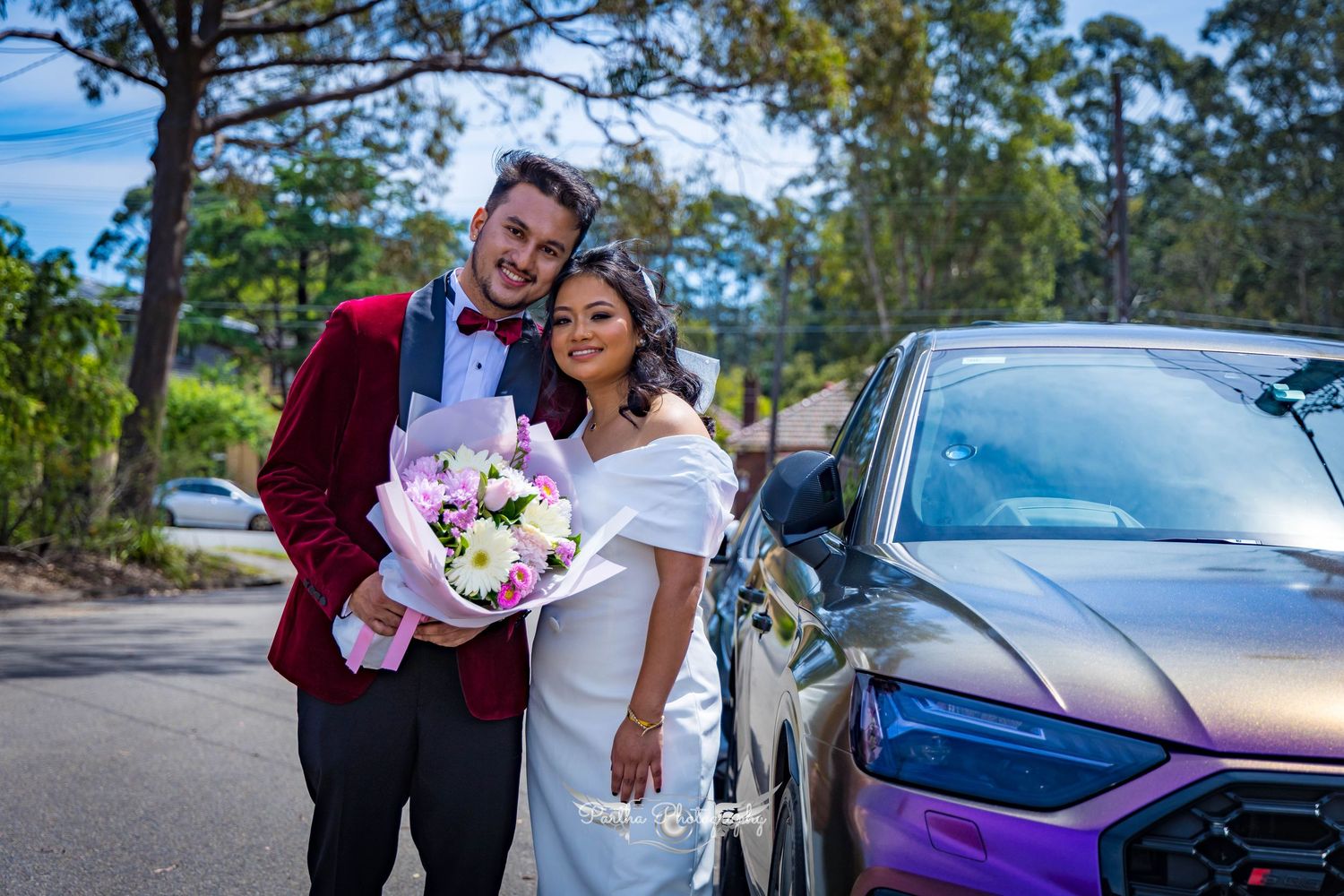 Couple in wedding attire poses with bouquet next to a purple car.
