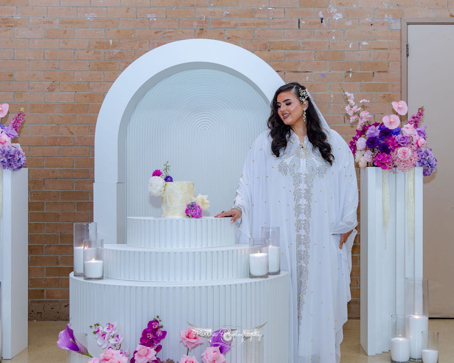 Wedding setup with white arch, candles, and pink and purple floral arrangements against a brick wall.