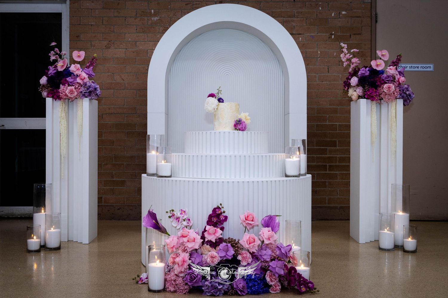White wedding cake decorated with pink and purple flowers displayed in an arched alcove.
