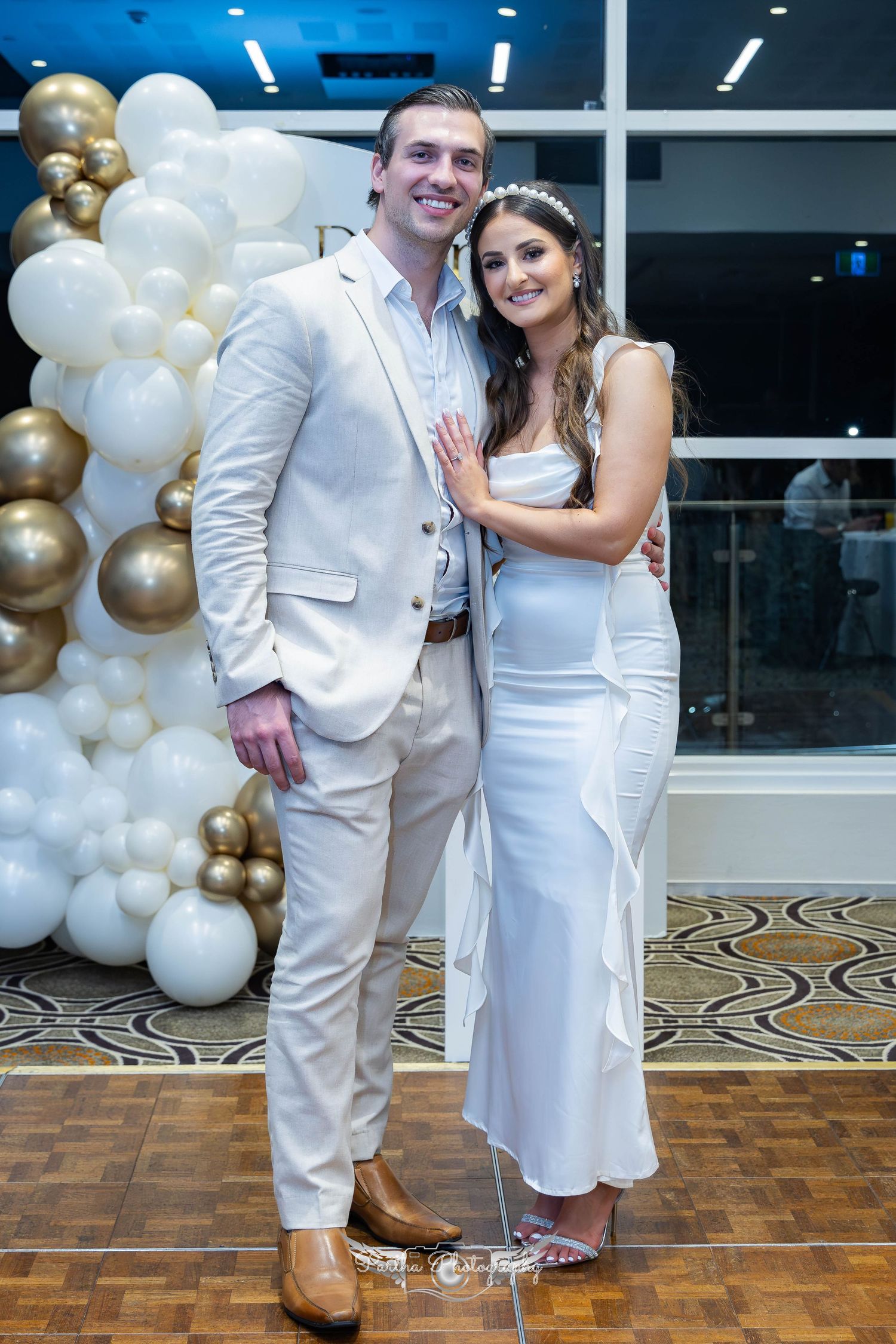 Newlyweds pose together against white and gold balloon backdrop at reception.