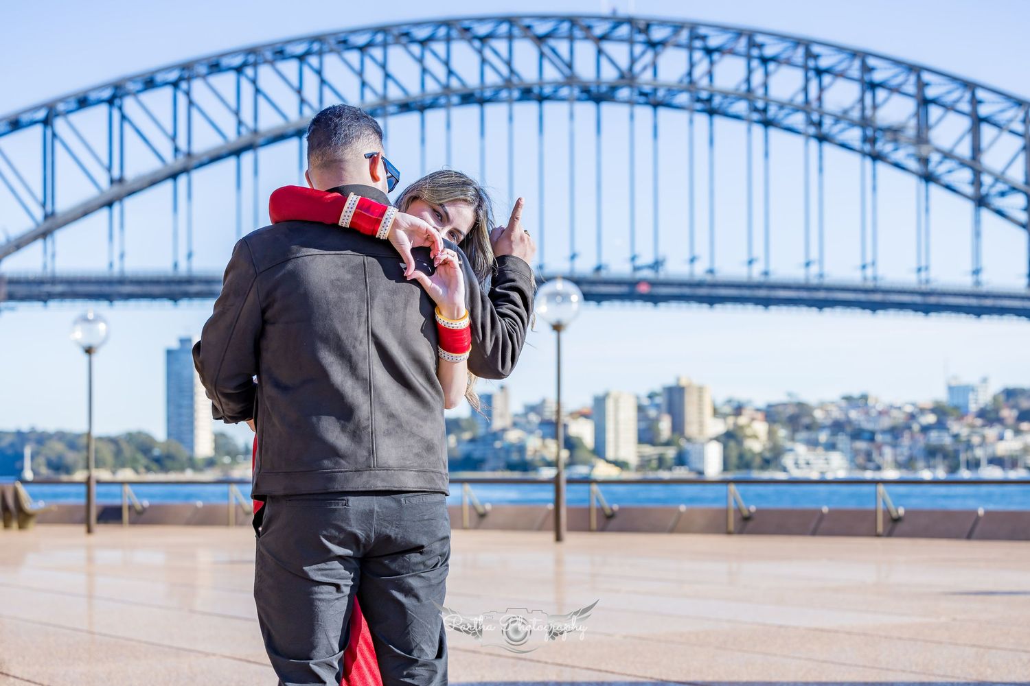 A couple embraces along Sydney Harbour with the iconic Harbour Bridge in the background on a sunny day.