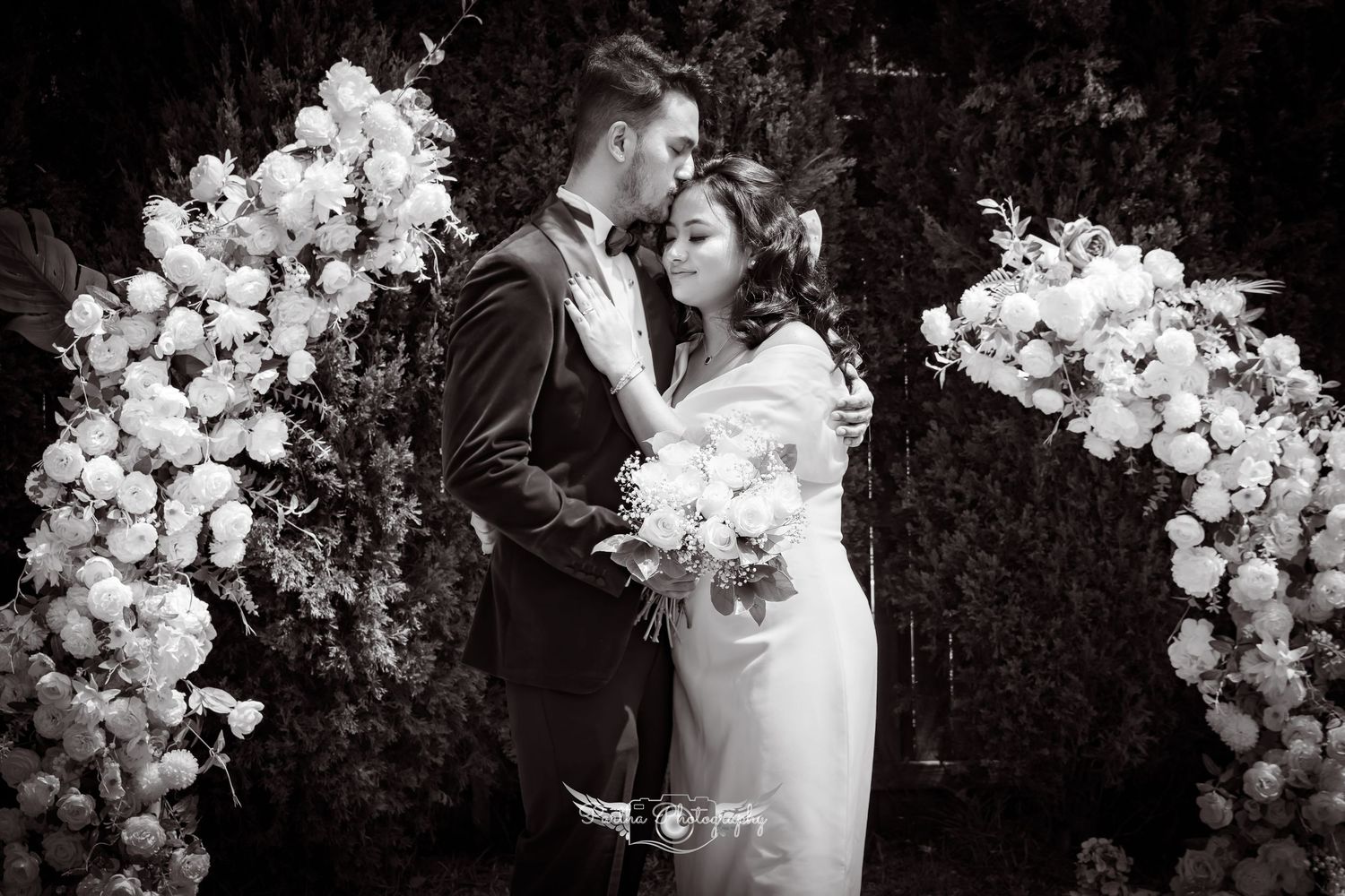Black and white romantic portrait of couple embracing under floral wedding arch.