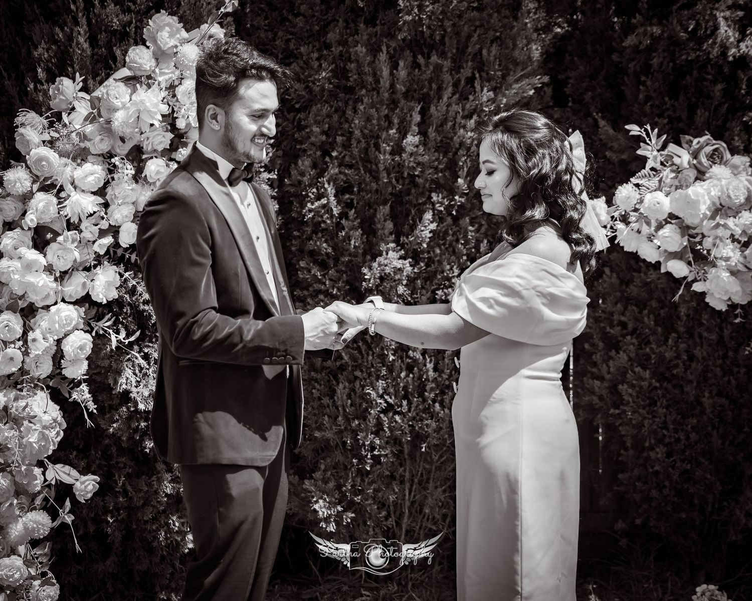 A black and white ceremony photo of a couple exchanging rings under an elegant floral arch.
