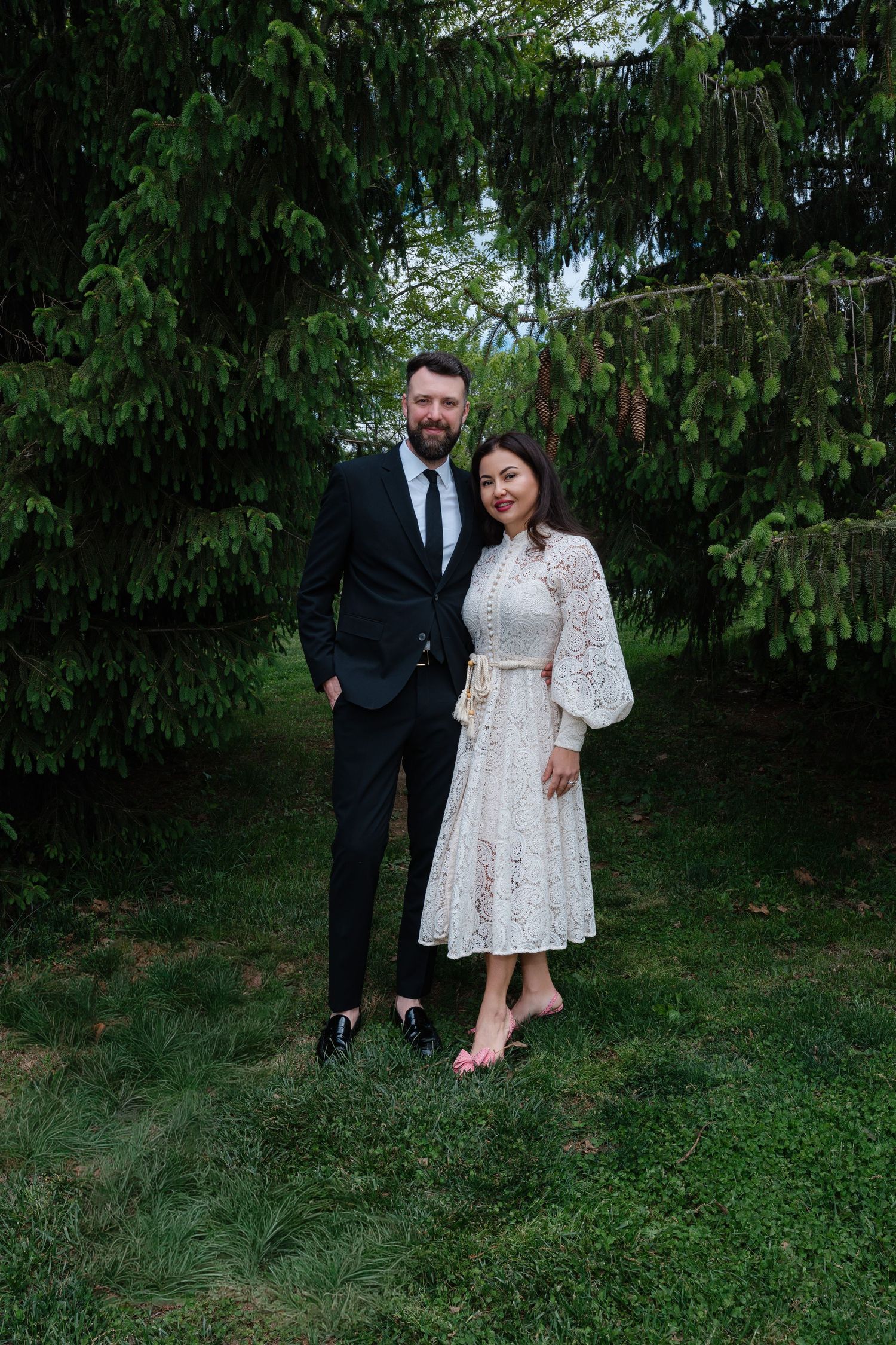 Couple posing outdoors in formal attire, man in black suit and woman in white lace dress, surrounded by green trees.
