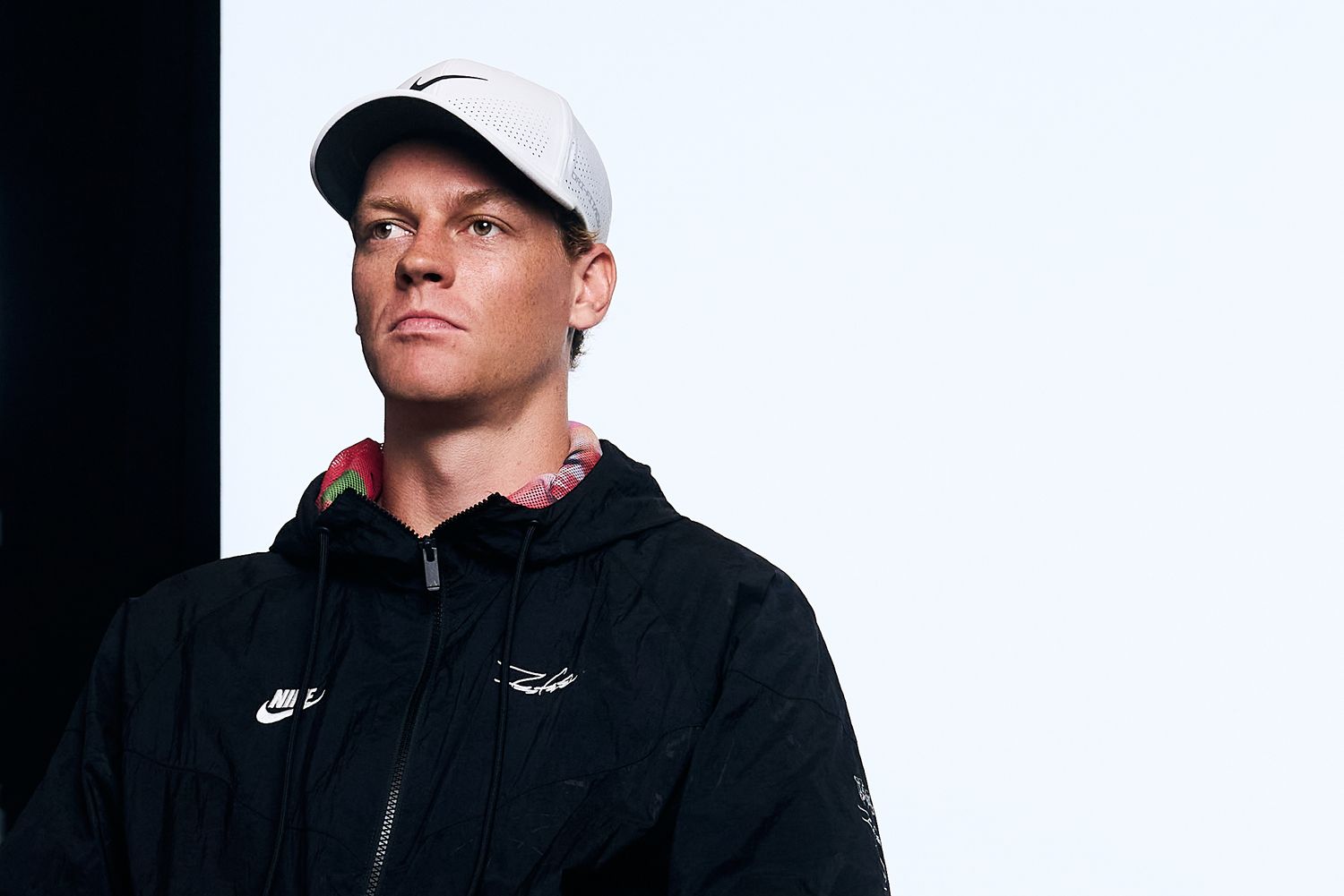 Professional athlete in black Nike jacket and white baseball cap poses for portrait series against dark background.
