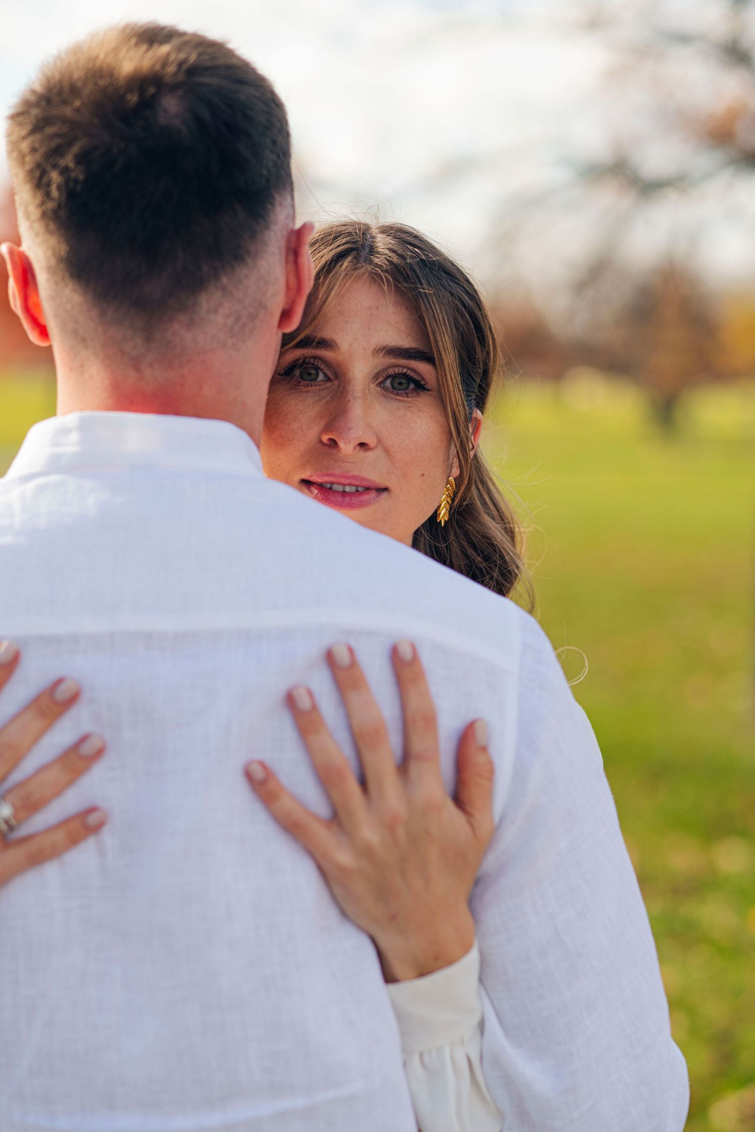 A romantic couple embraces outdoors with the person in white clothing facing away from the camera in a natural setting.