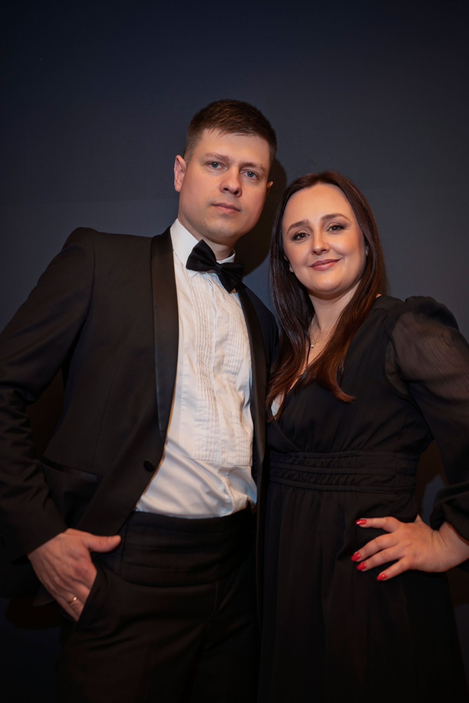 A couple in formal wear poses intimately against a dark background.
