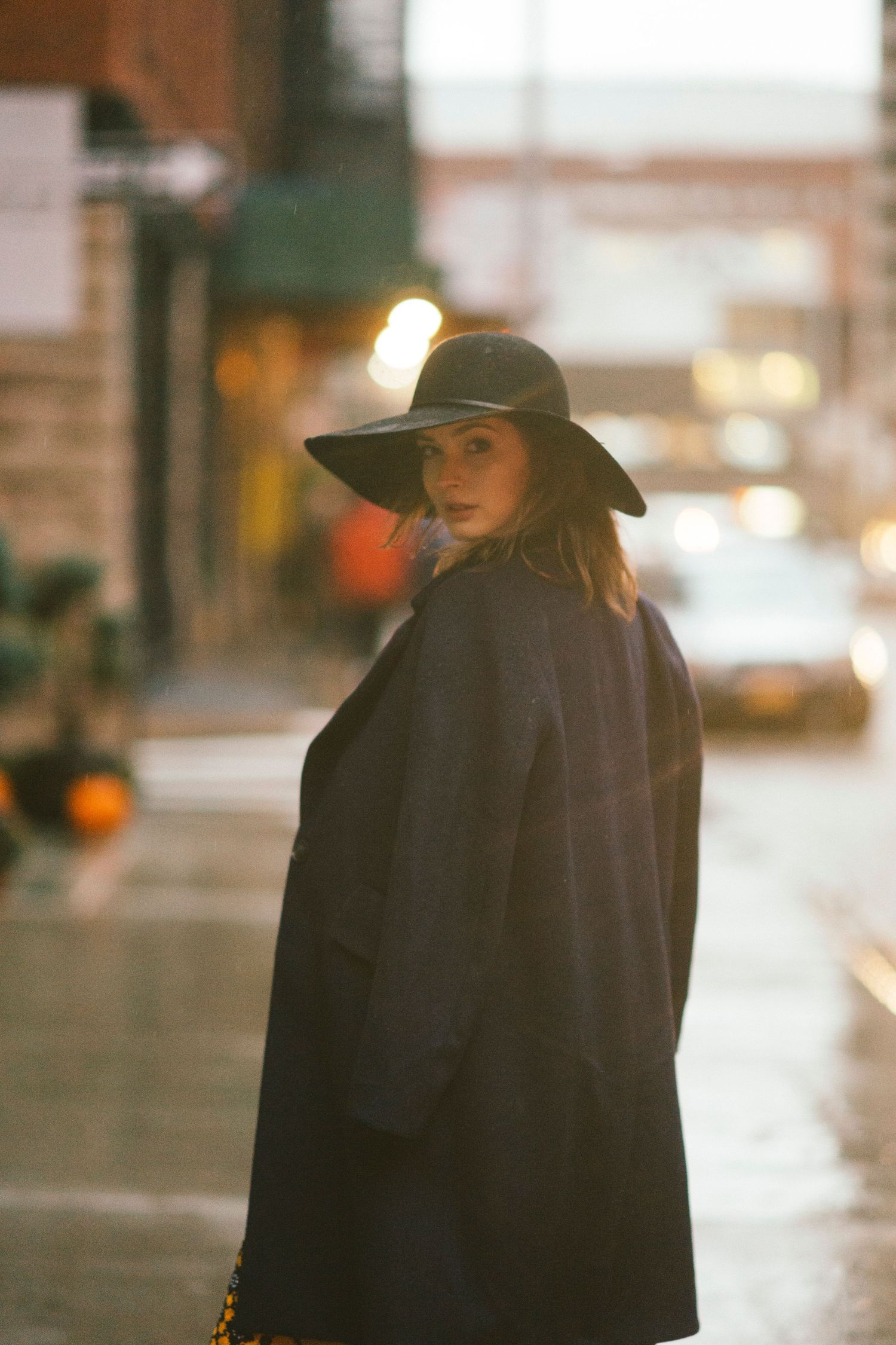 A person in a black coat and wide-brimmed hat poses on a rainy city street at dusk.