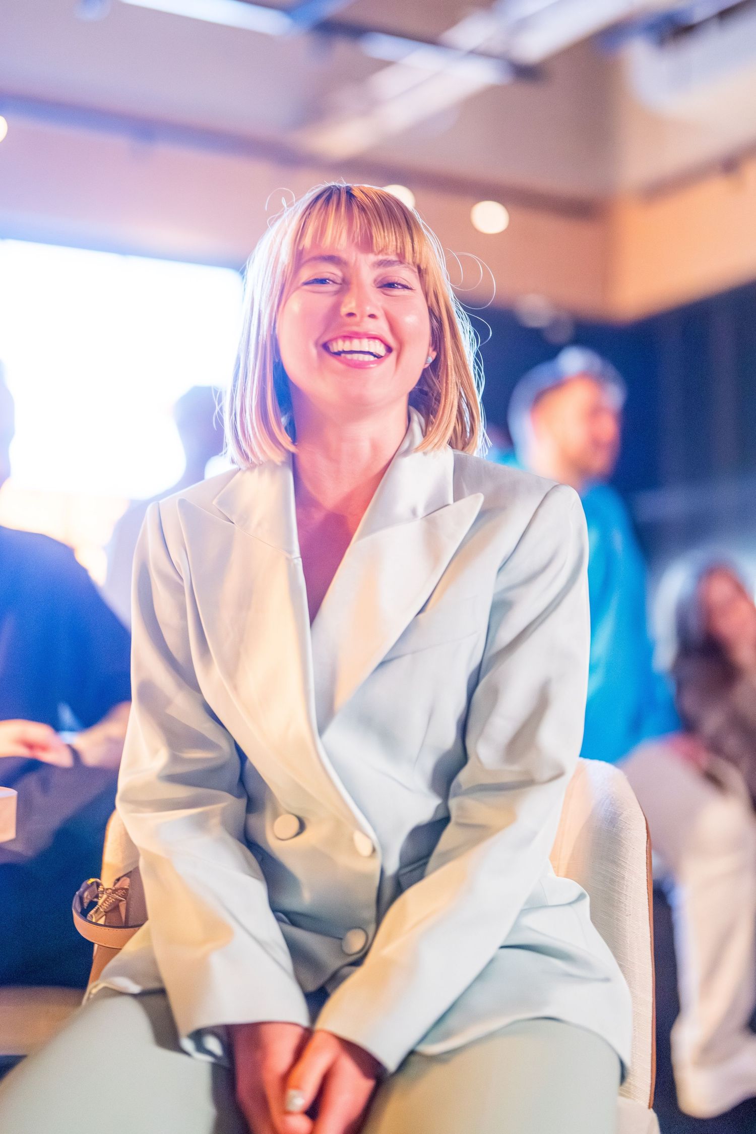 Smiling woman in a light blue blazer suit sitting at an indoor event, laughing joyfully.