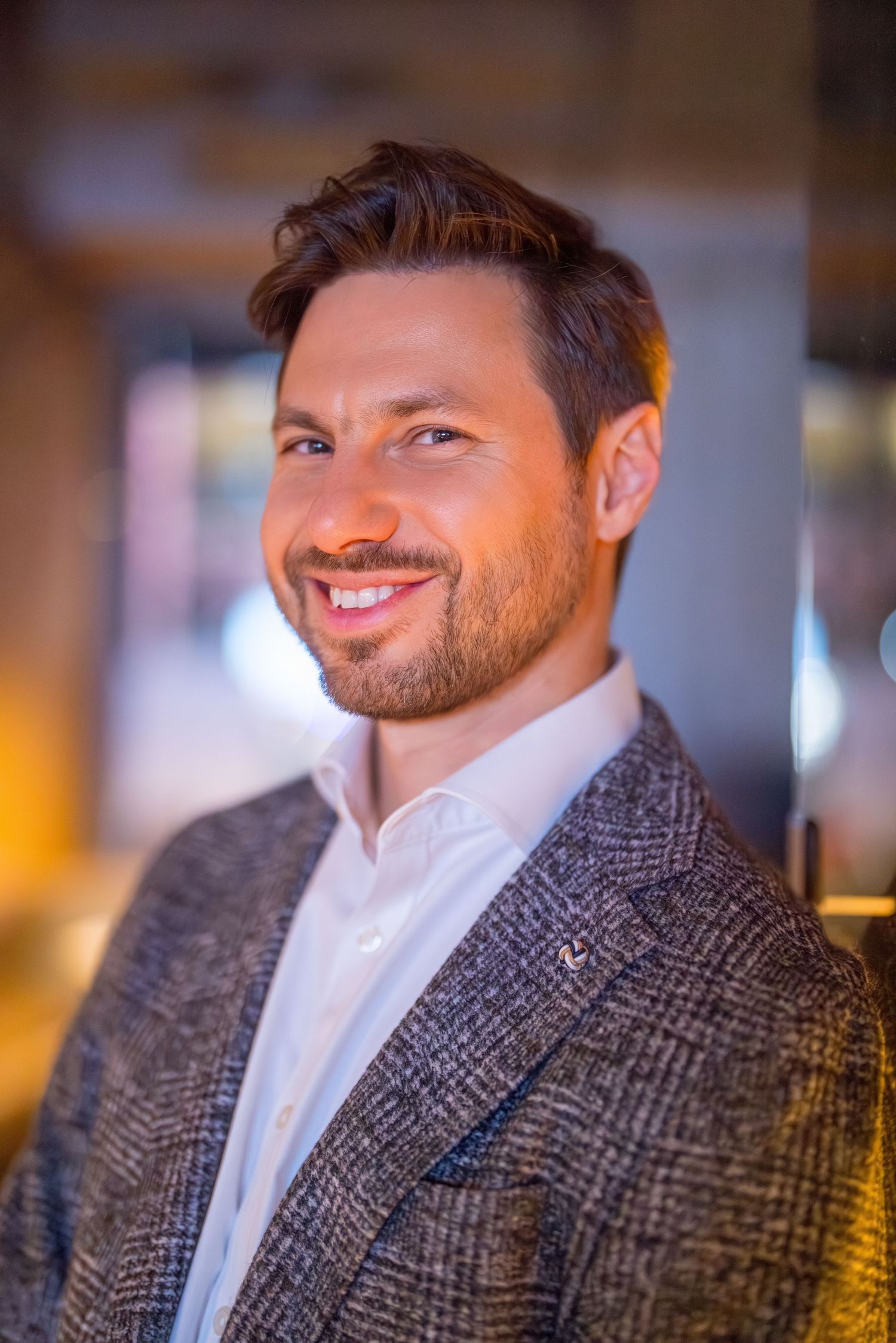 Smiling man with beard wearing a gray tweed blazer and white shirt in a warmly lit indoor setting.