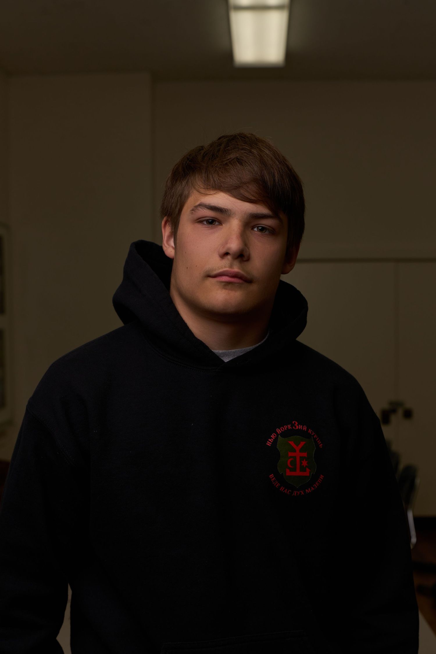 Young man in black hoodie with red logo poses seriously indoors under dramatic lighting.