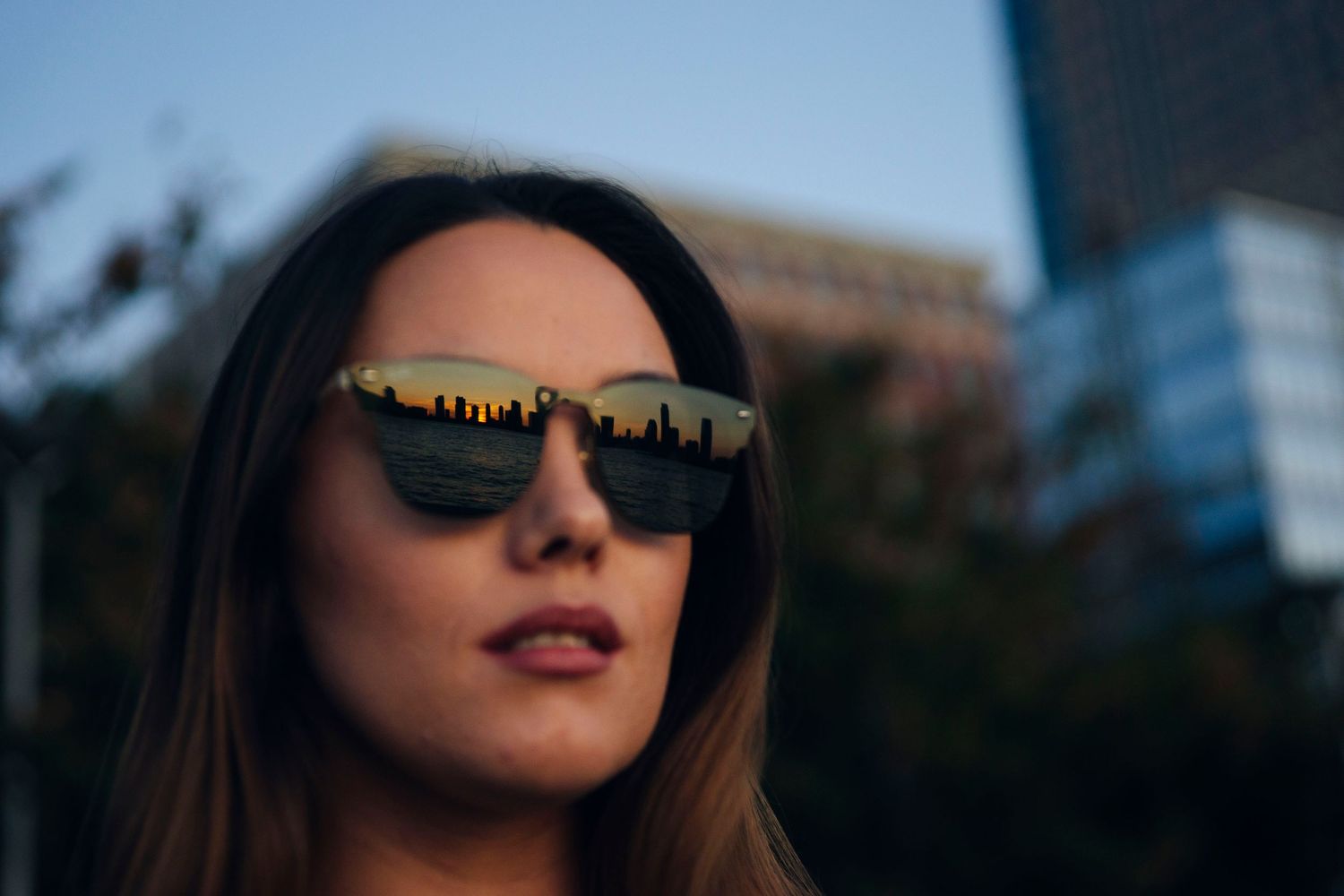 Stylish portrait with city skyline reflection in sunglasses at dusk.