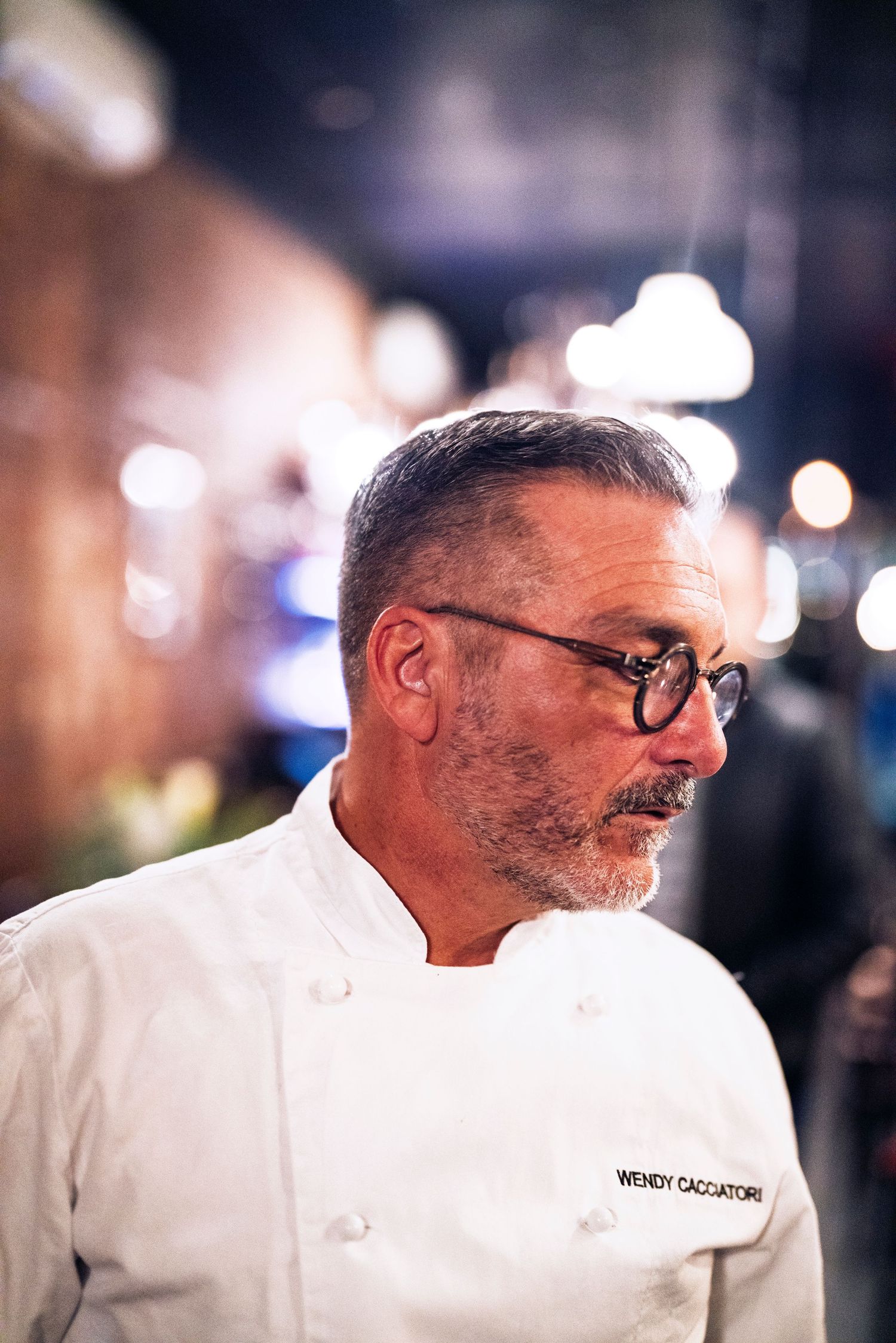 A chef in white uniform and glasses stands in profile against a blurred nighttime background with bokeh lights.