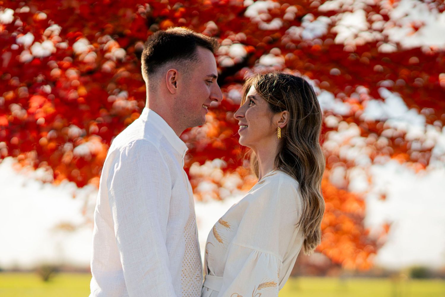 A couple in white clothing stands close together against red fall foliage.