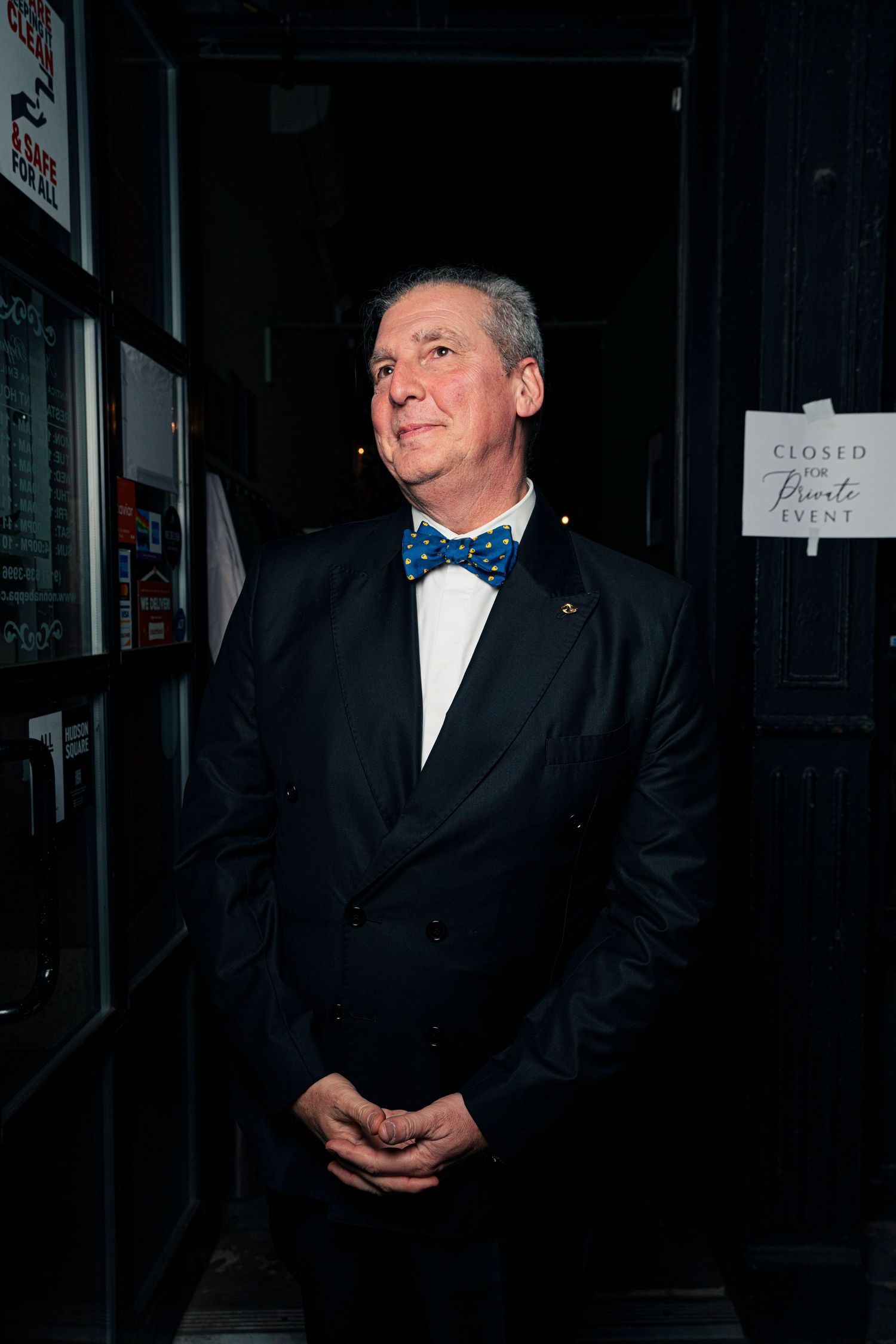 A person in a dark suit smiles while standing against a black background in a dimly lit setting.