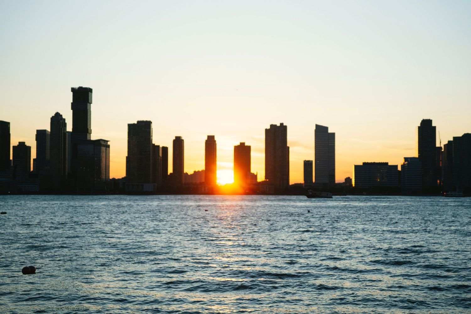 Dramatic city skyline silhouette at sunset with sun peeking between skyscrapers.