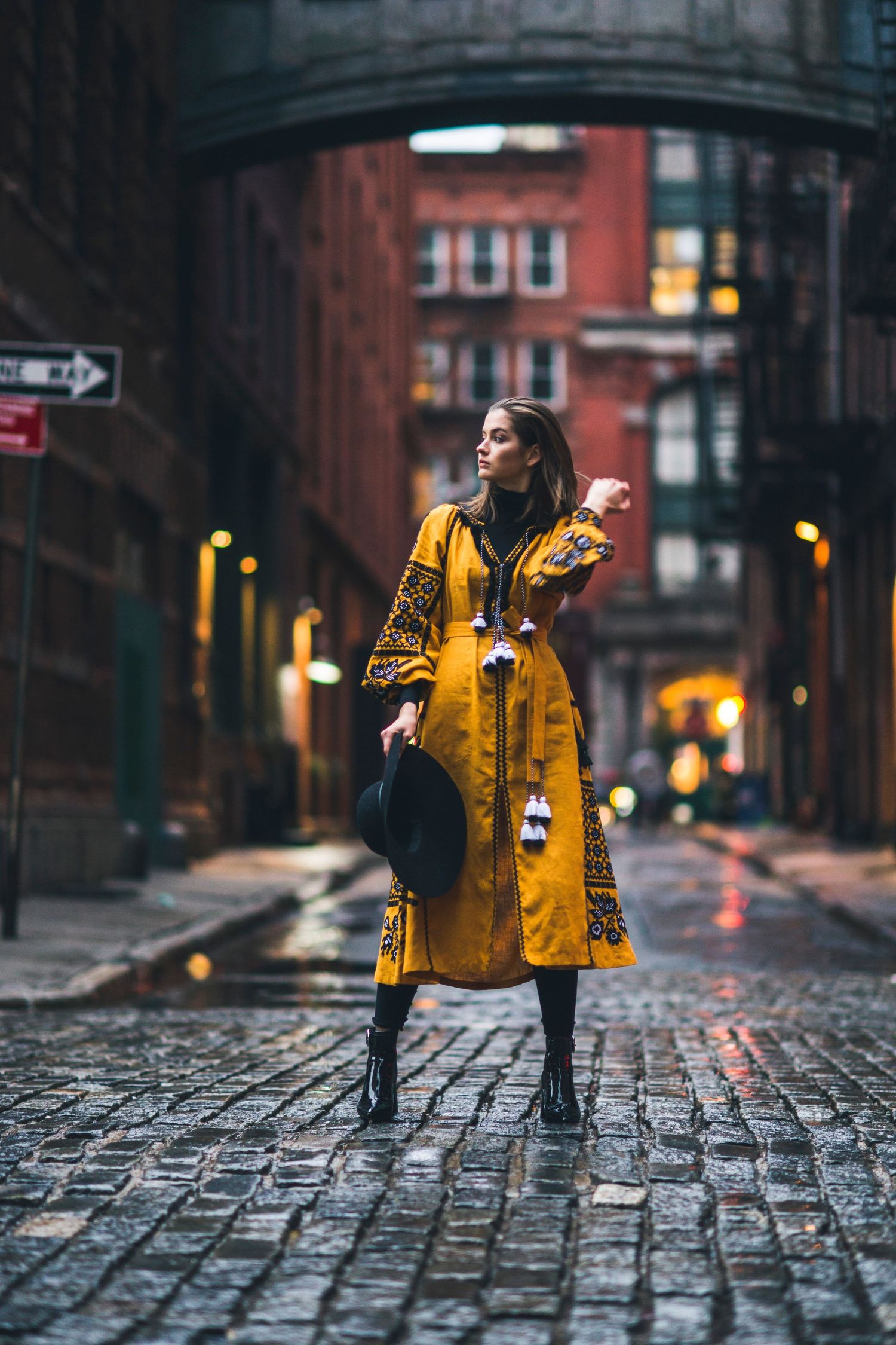A person wearing a yellow patterned coat poses on wet cobblestone streets in an urban setting.