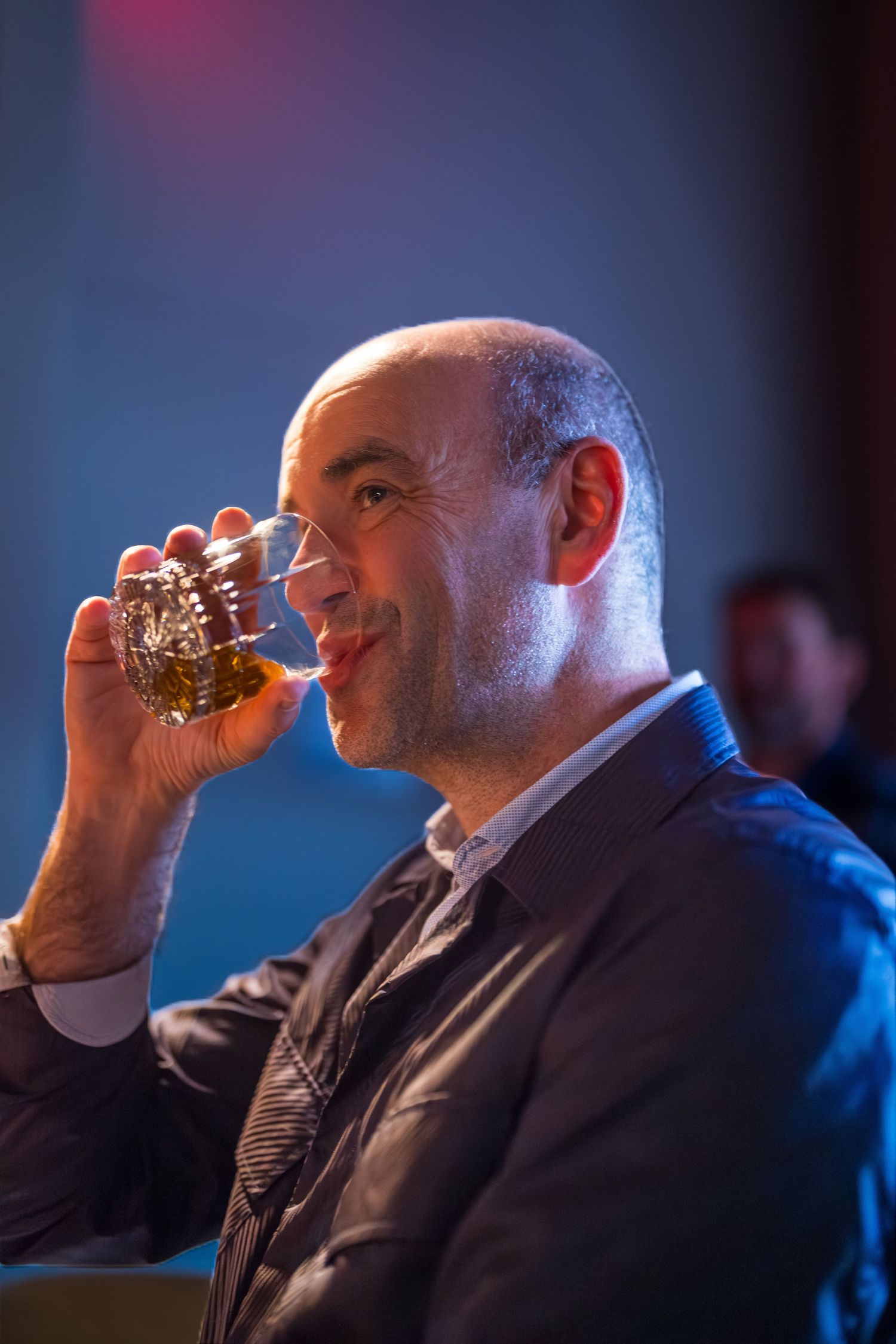 Bald man in a suit sipping whiskey from a glass in a dimly lit venue.