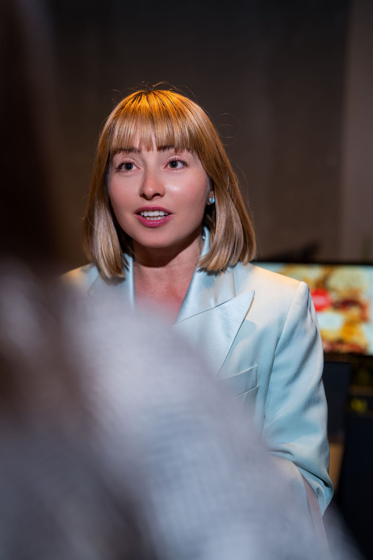 Woman with blonde bob haircut in light blue blazer speaking at an indoor event.