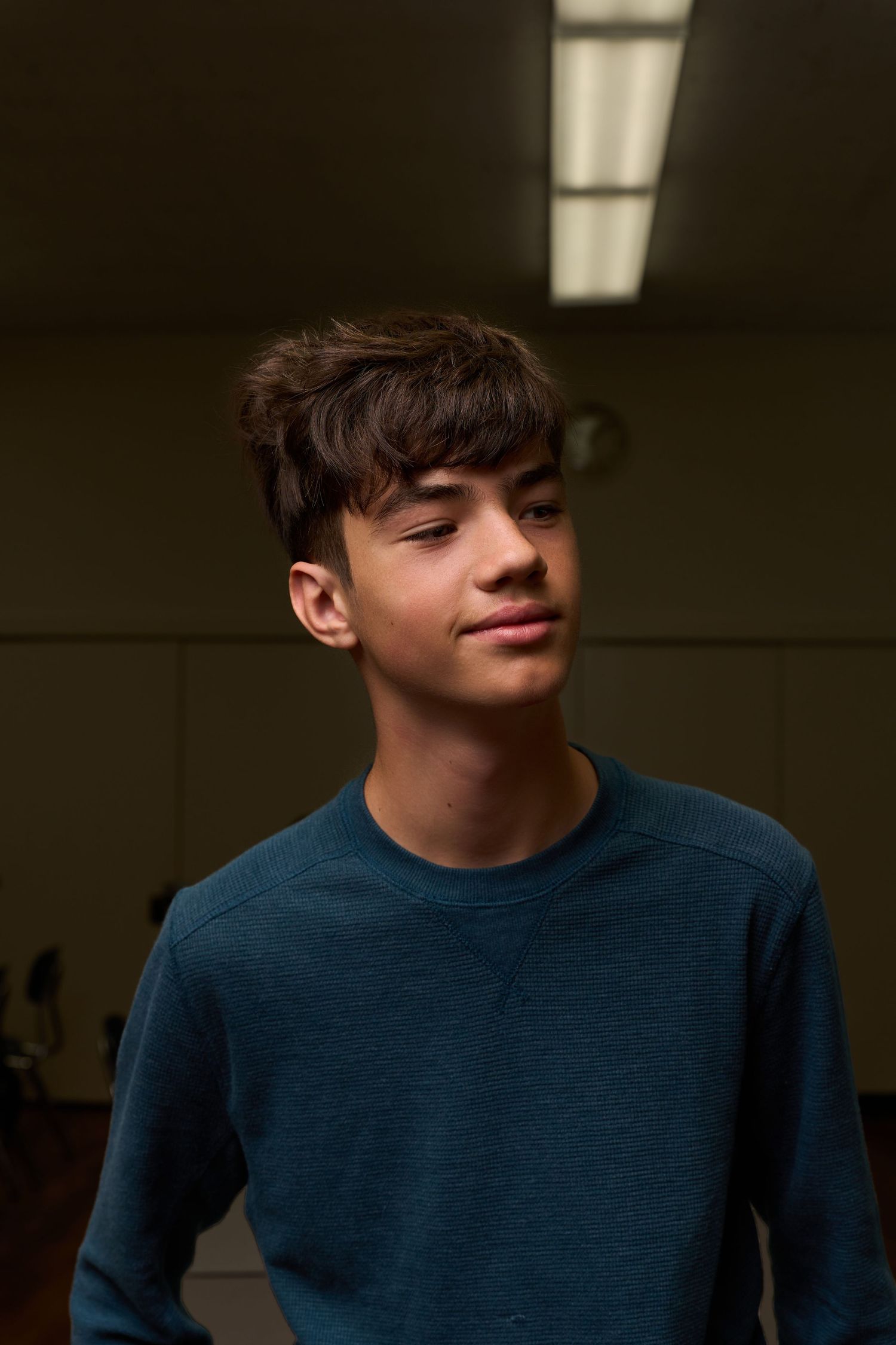 A young teen boy with dark hair wearing a teal sweatshirt stands in a dimly lit room.