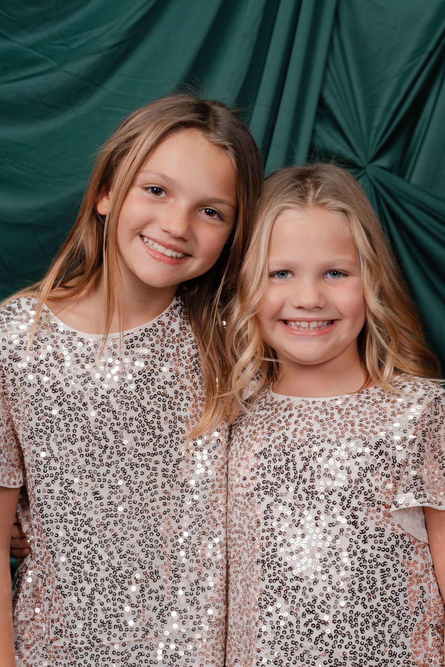 Two smiling girls in matching sequined dresses pose together in front of a green draped backdrop.