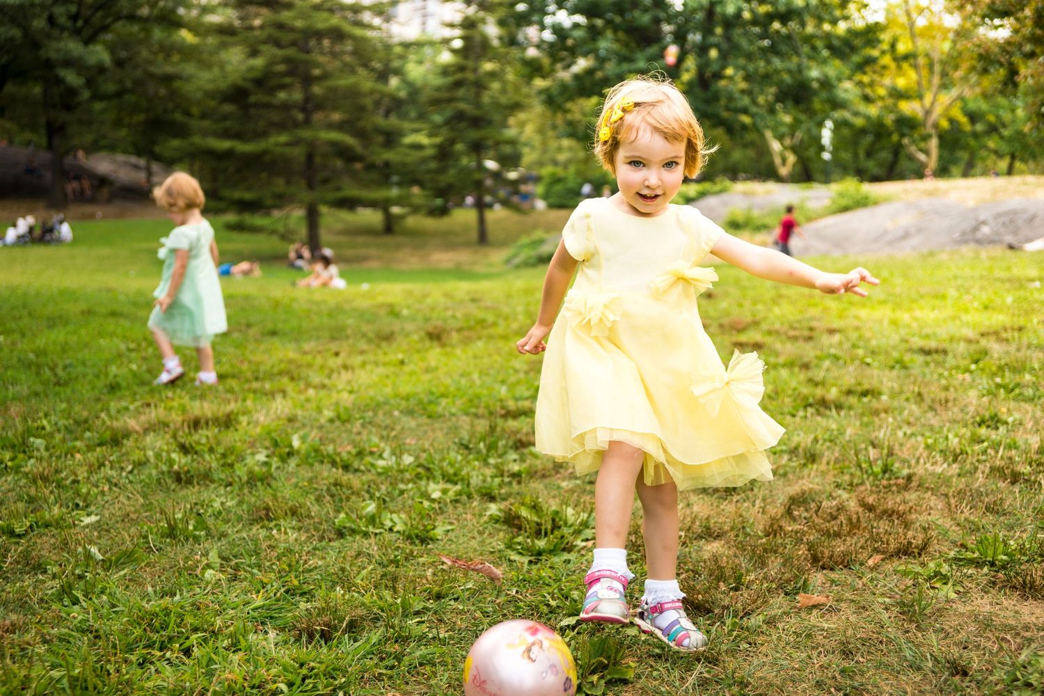 A child in a yellow dress plays on a grassy hill with a ball on a sunny day.