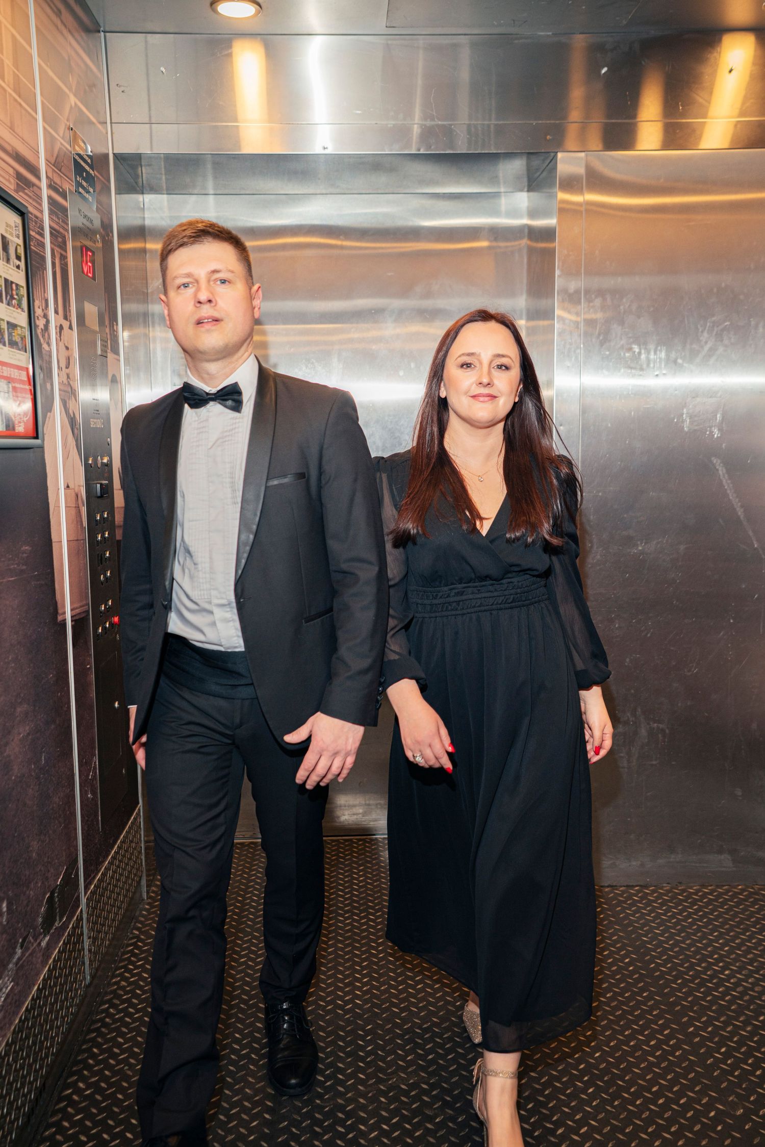 A couple in black formal attire poses together in an industrial elevator.