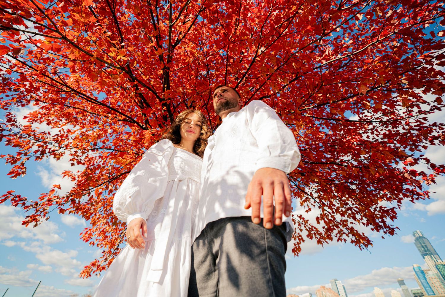 A couple poses under vibrant red autumn maple leaves against a blue sky.
