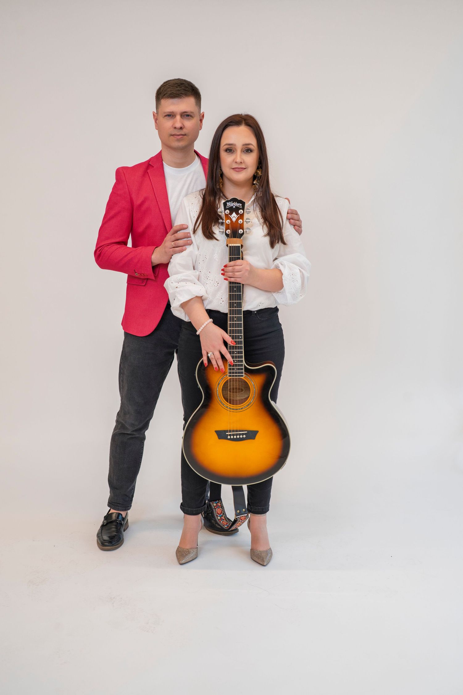 Duo poses with acoustic guitar against white background in casual attire.