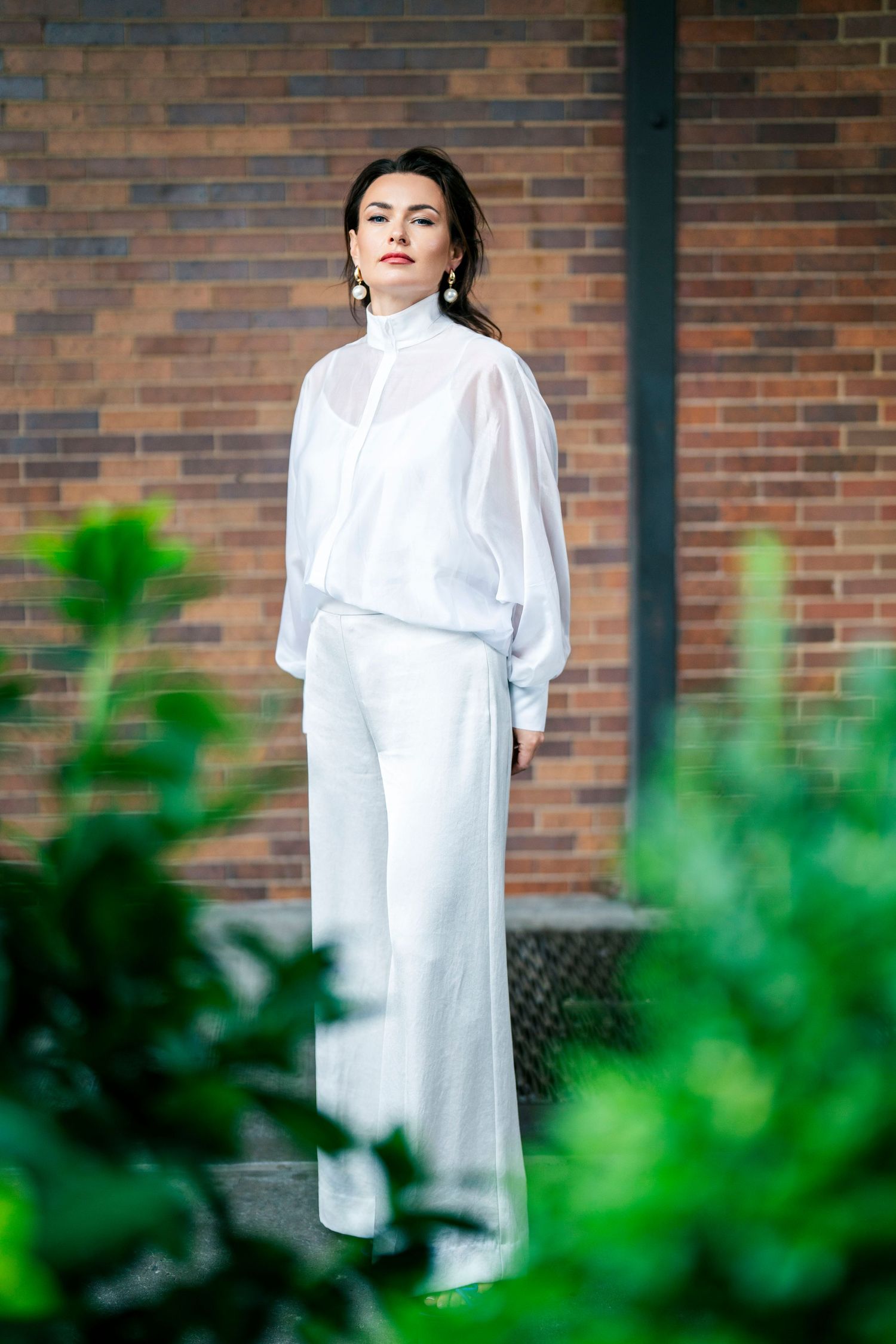Fashion model in white outfit poses against brick wall with green foliage in foreground.