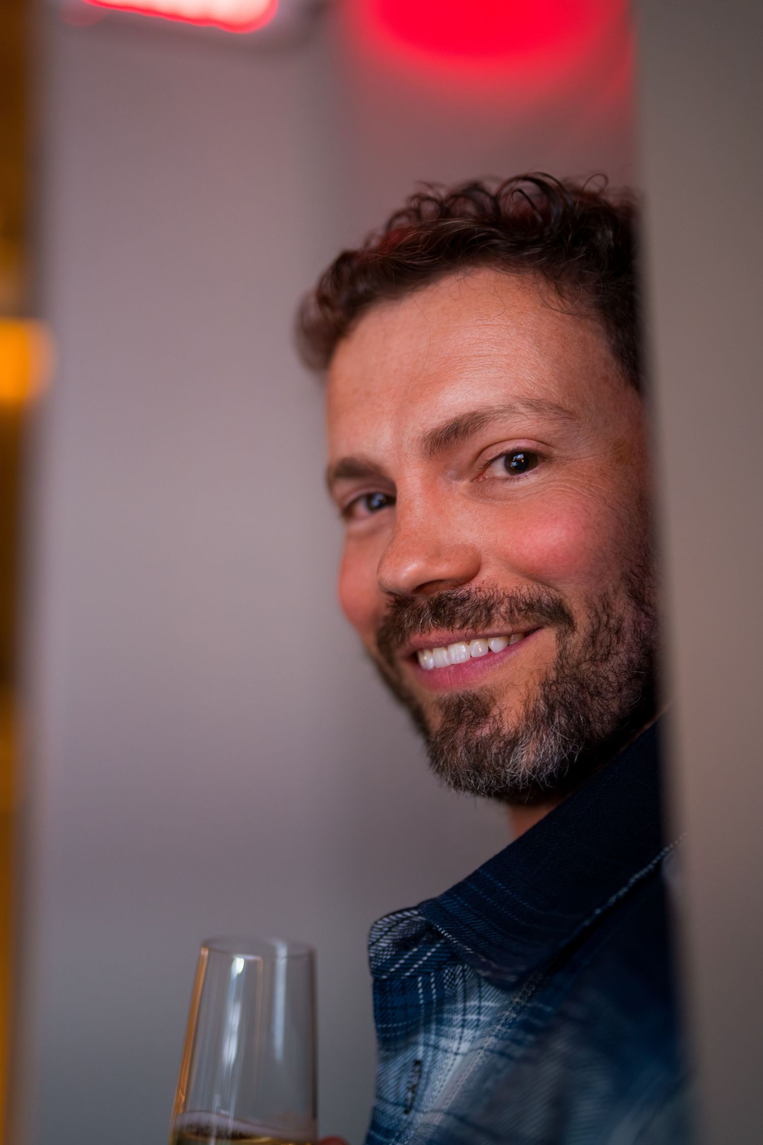 Smiling man with beard holding champagne glass peeking around corner in dimly lit bar.