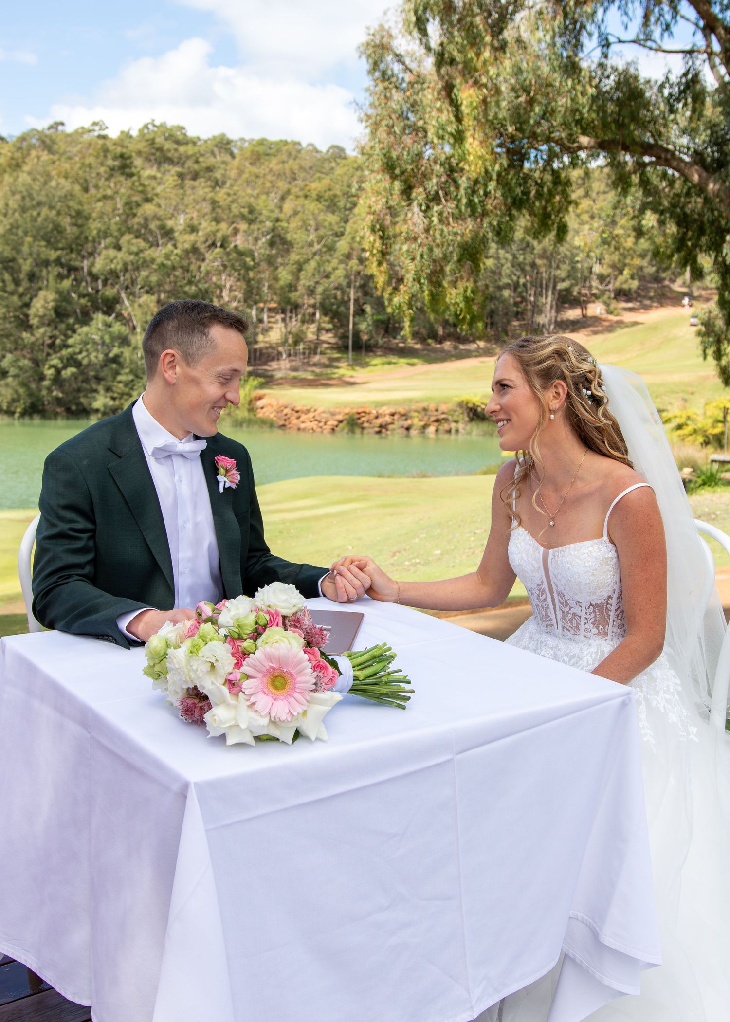 Wedding couple sits together at a white-clothed table outdoors on a golf course with trees in the background.