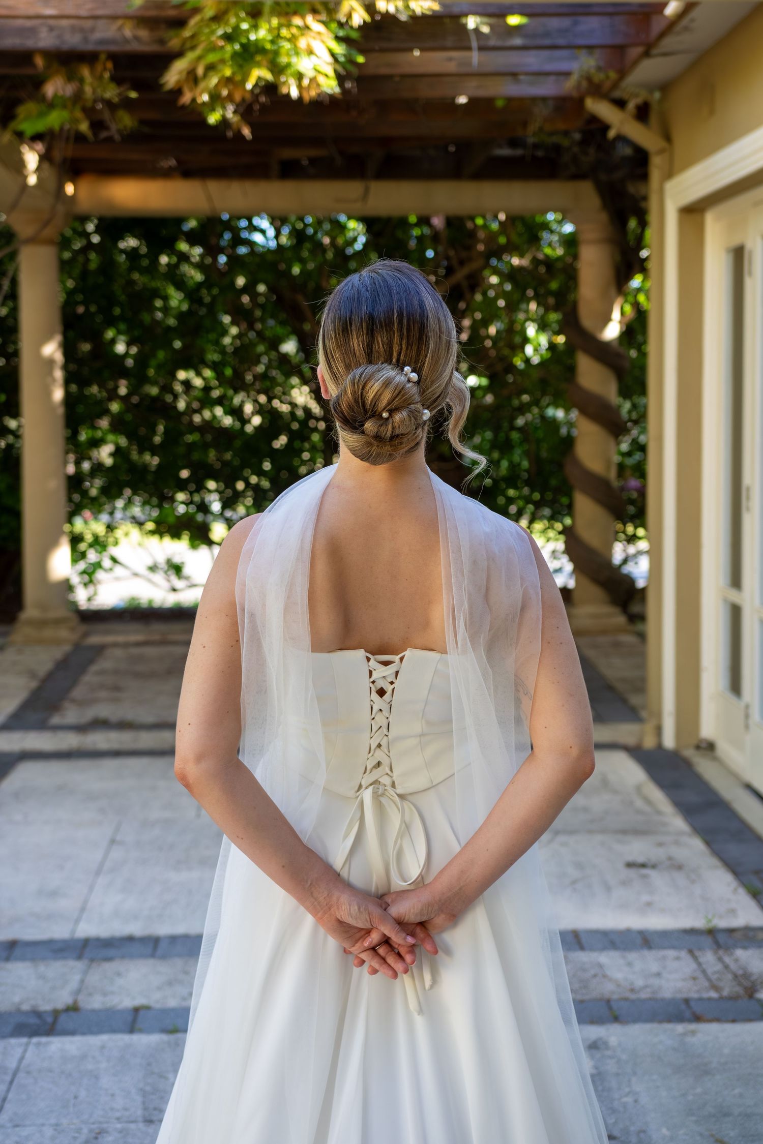 Back view of a bride in a white corset wedding dress with lace-up detail standing under an outdoor pergola.