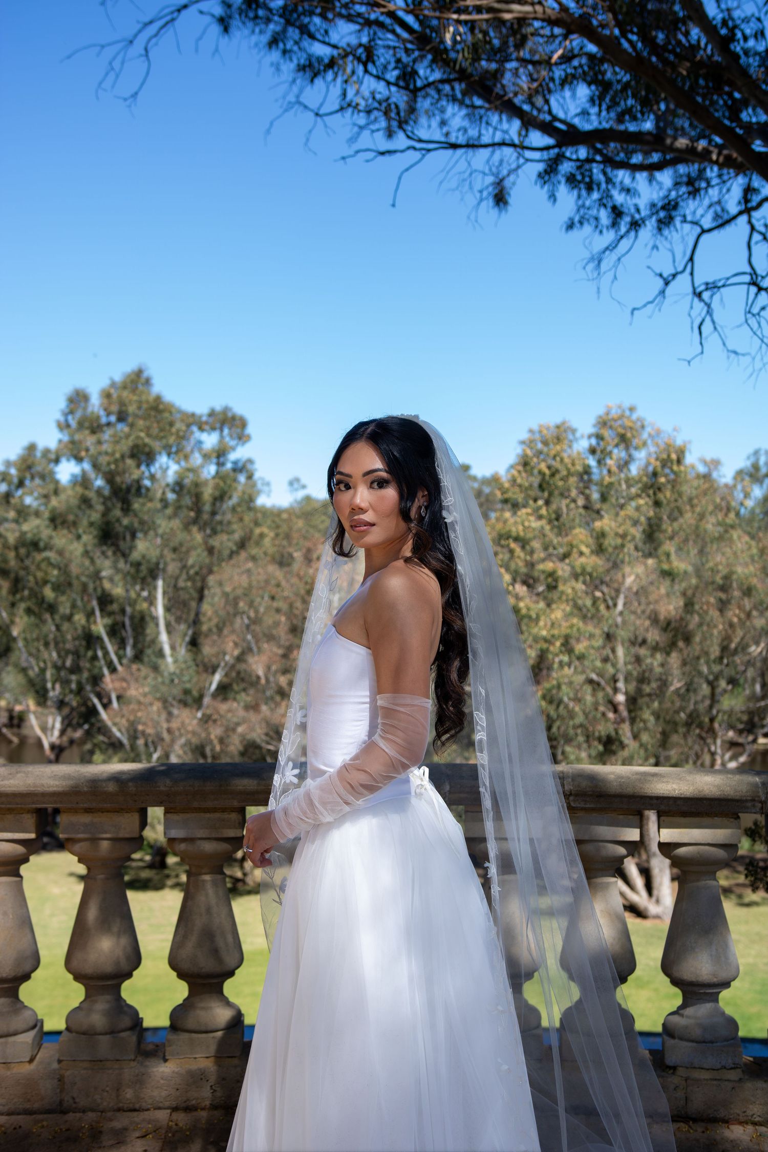 A bride in a white strapless wedding gown and long veil poses on a stone balcony with trees in the background.