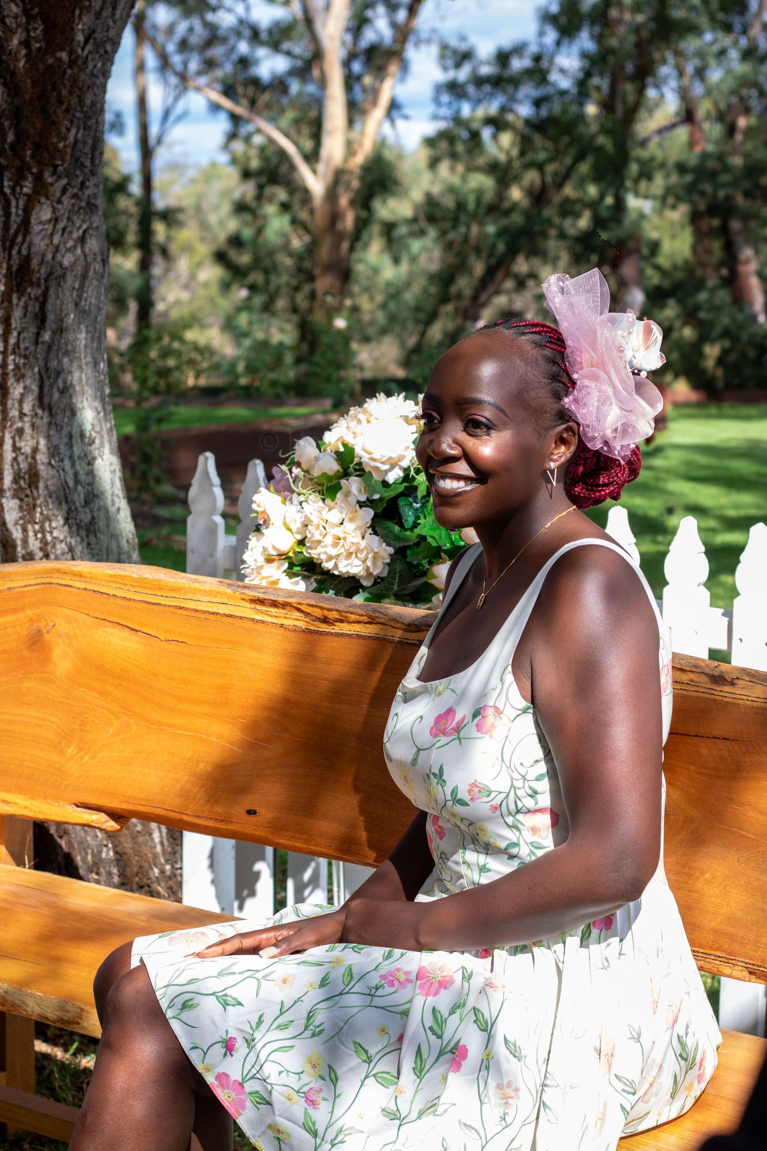 Outdoor portrait showing a floral dress and pink hair accessory against a wooden bench setting in a sunlit garden.