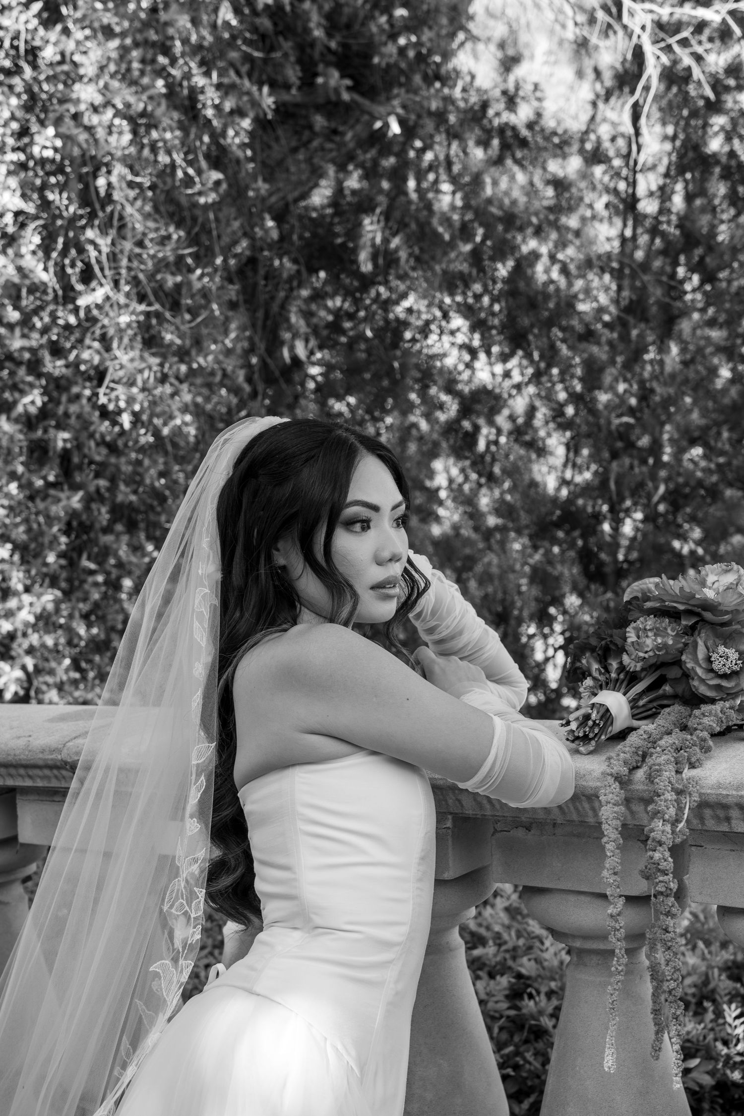 A black and white wedding portrait showing a bride with flowing veil in a strapless gown against a natural backdrop.