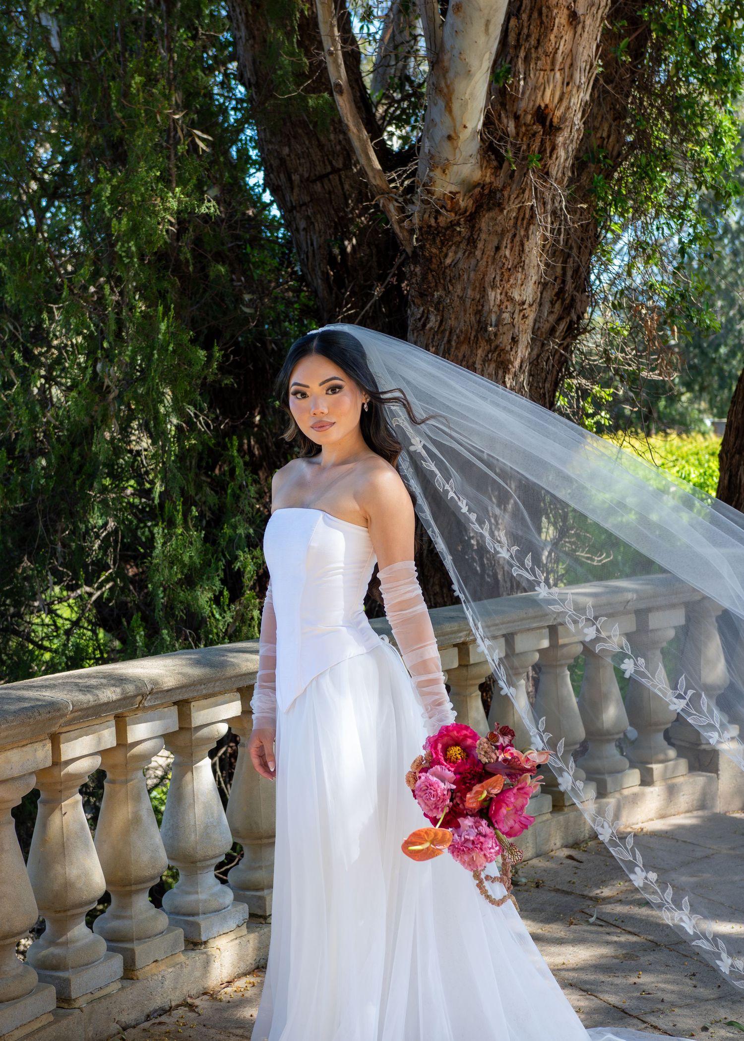 A bride in a flowing white gown holds a pink bouquet while posing along an elegant stone balustrade in a garden setting.