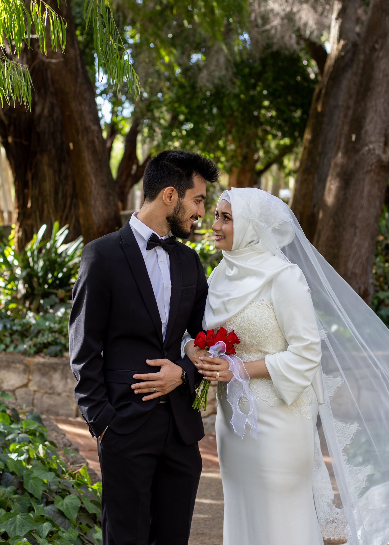 Muslim bride in white hijab and groom in black tuxedo exchanging smiles in a lush garden wedding photo.