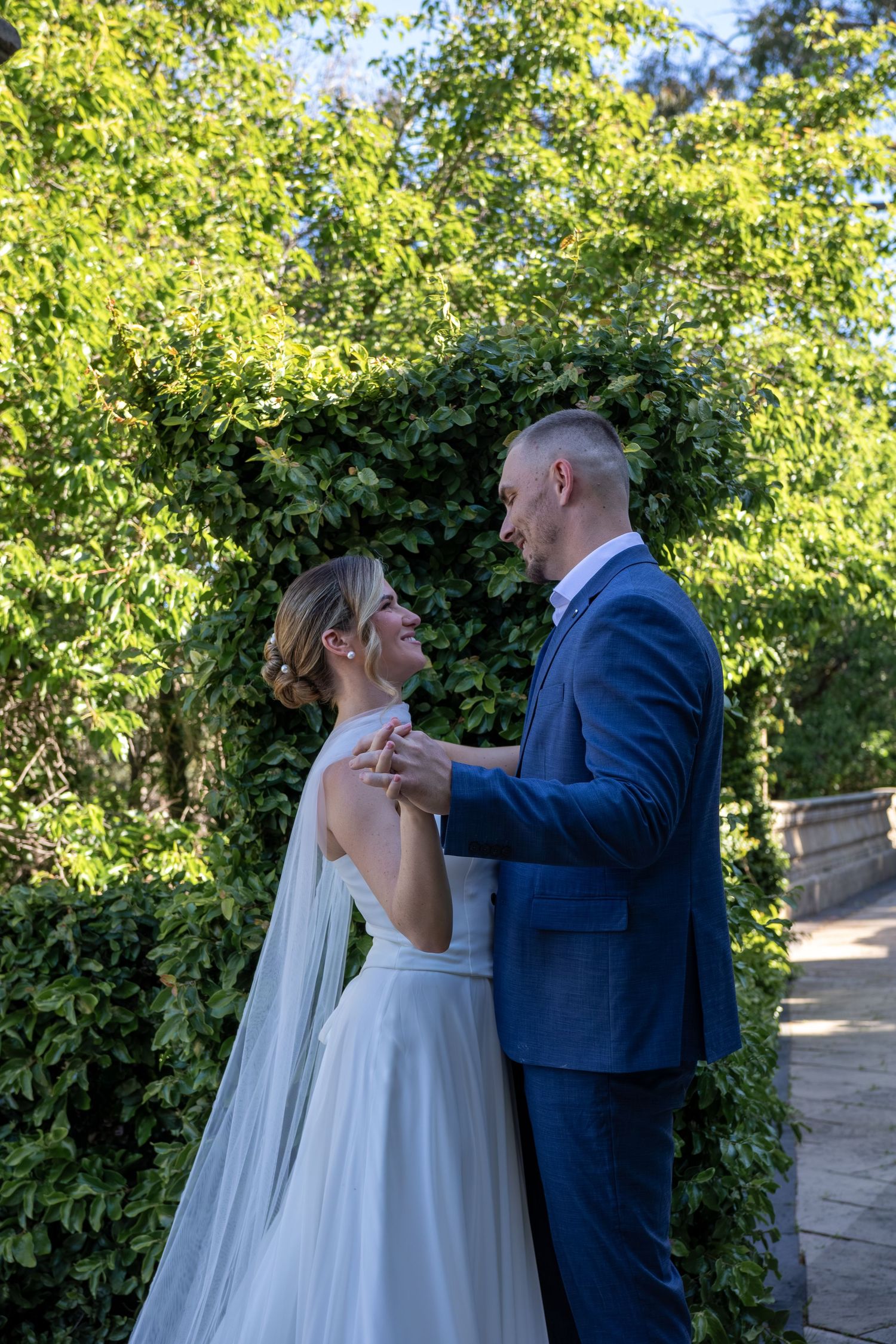 A couple shares an intimate moment in a garden setting with lush greenery and sunlight filtering through the trees.