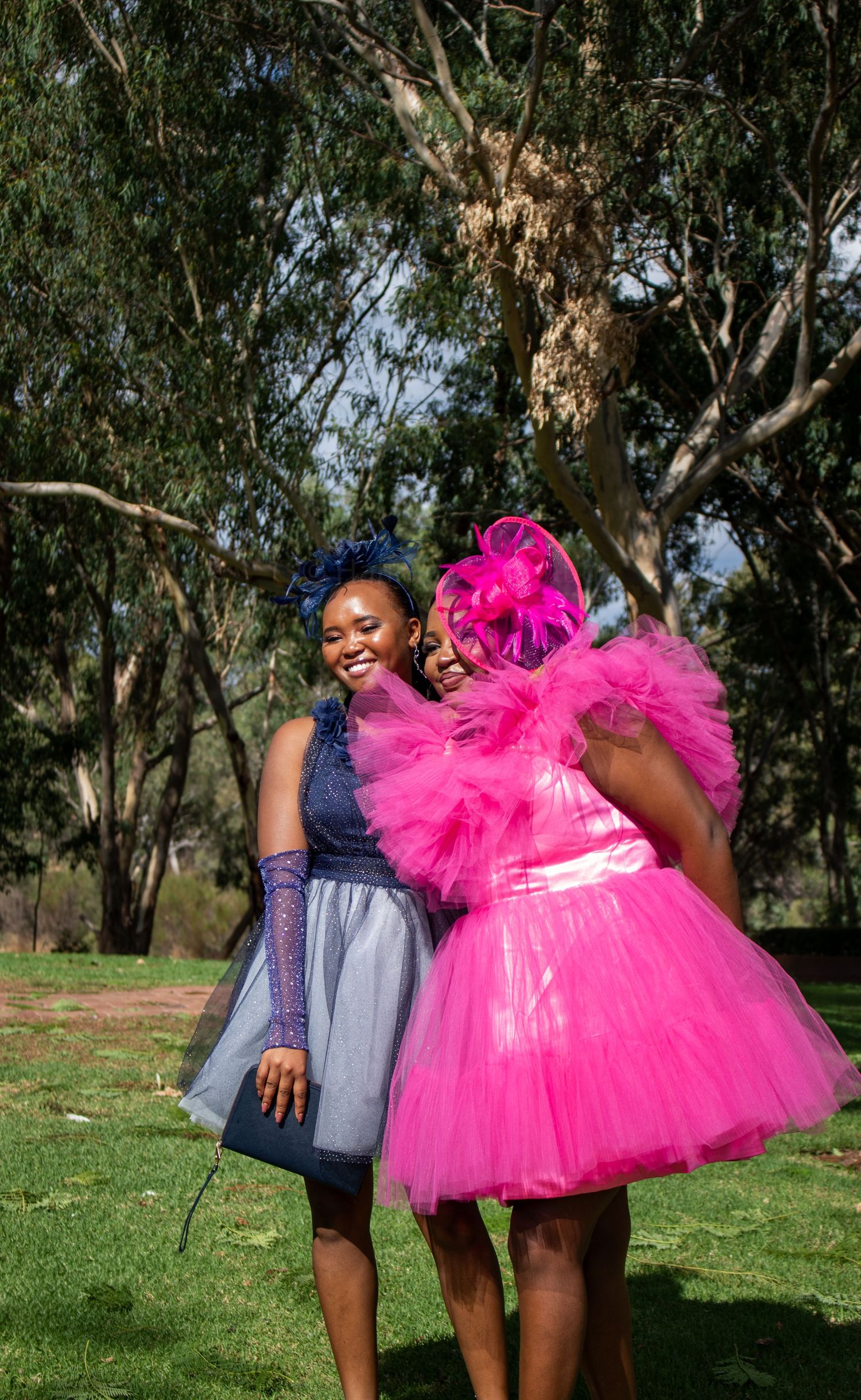 Two people in contrasting outfits, one in a navy dress and one in a bright pink tulle skirt, dancing joyfully in a park.