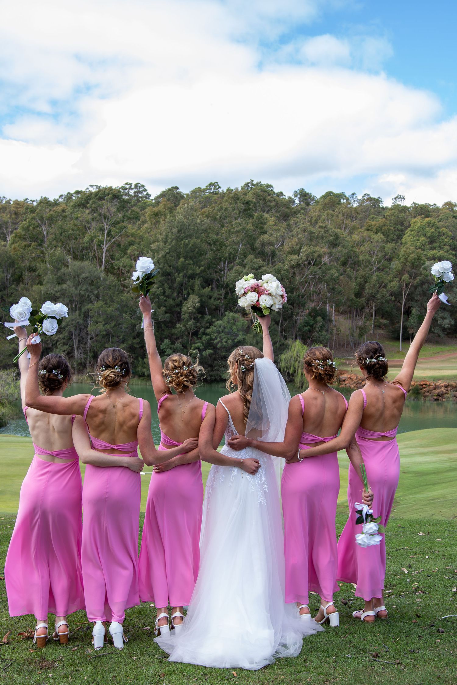Bride and bridesmaids in pink dresses pose with their backs turned holding bouquets on a green lawn with trees in background.