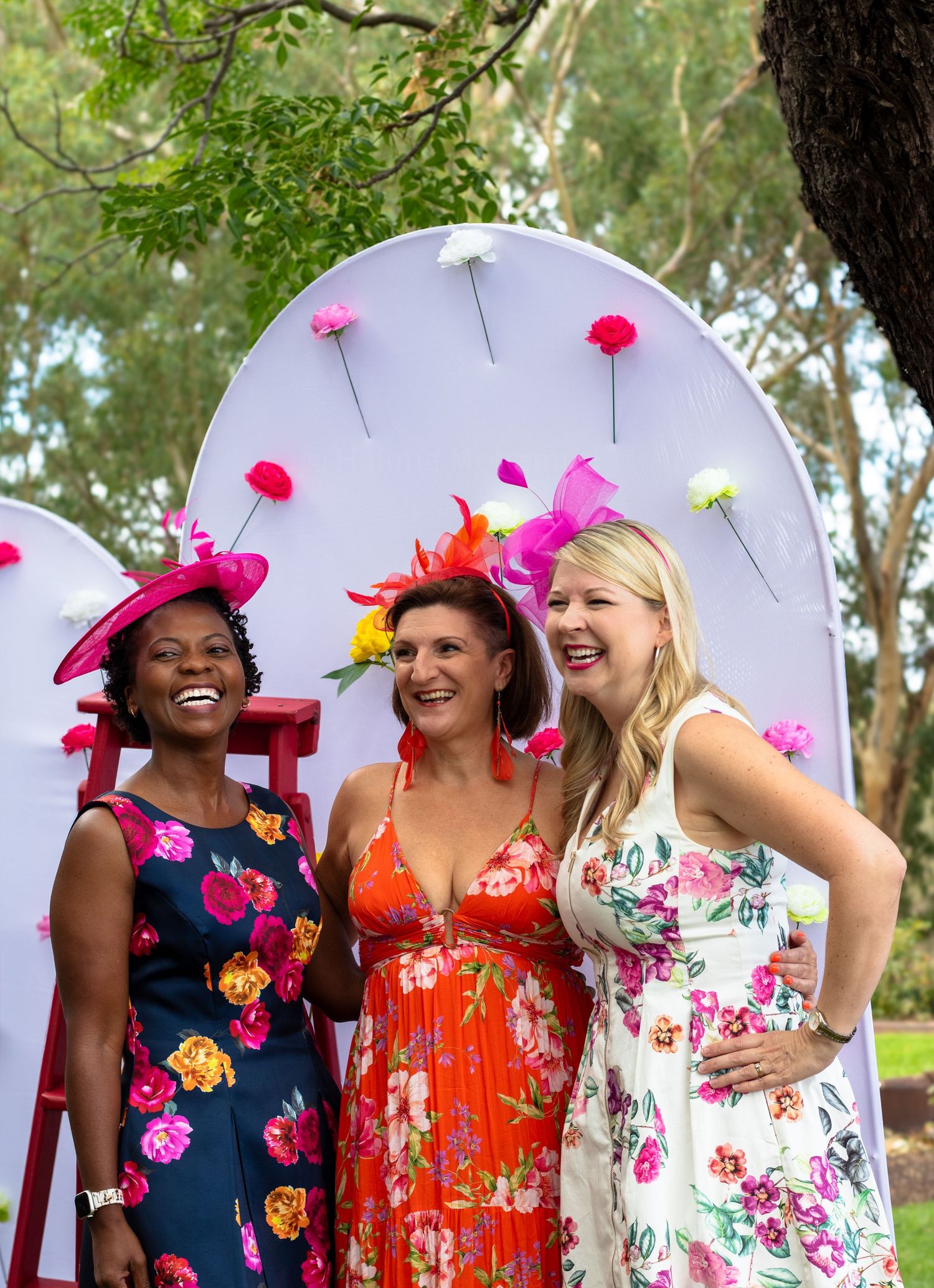 Three friends in colorful floral dresses and bright fascinators laugh together at an outdoor garden party.